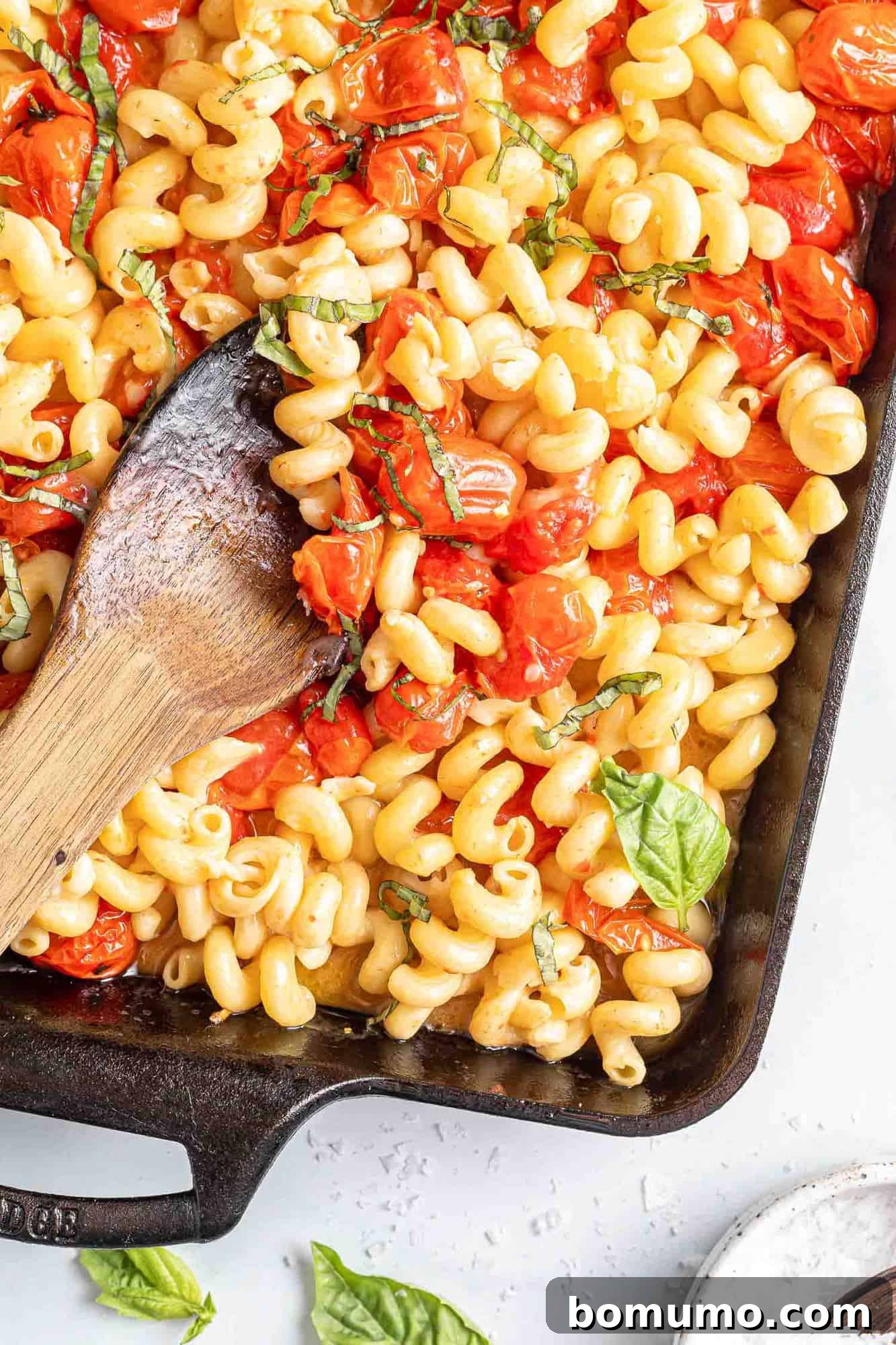 Pasta with vibrant cherry tomatoes, fresh basil, and a generous block of melted feta cheese in a black baking dish, ready to be tossed.