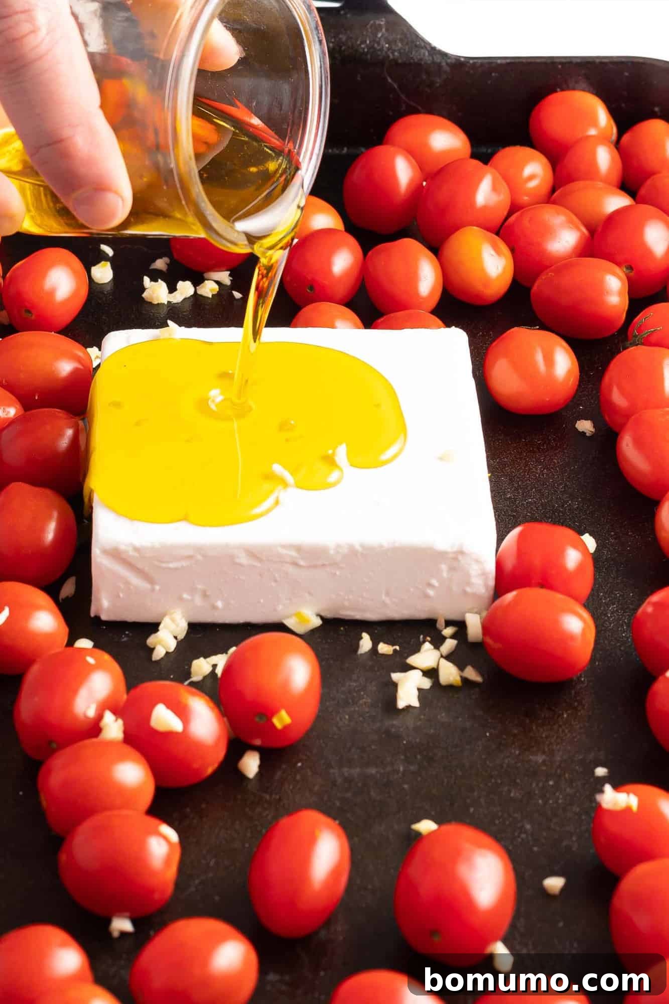 Golden olive oil being generously poured over a block of feta cheese and cherry tomatoes nestled in a baking dish, prepared for roasting.