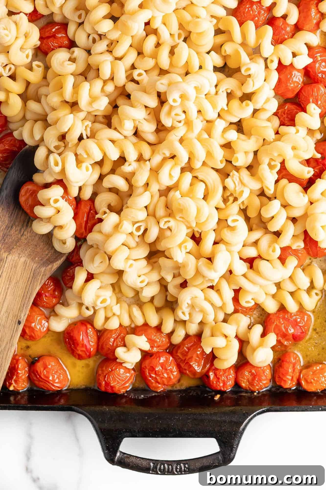 Cooked pasta being lovingly stirred into the vibrant roasted tomatoes and melted feta sauce within a black baking dish.