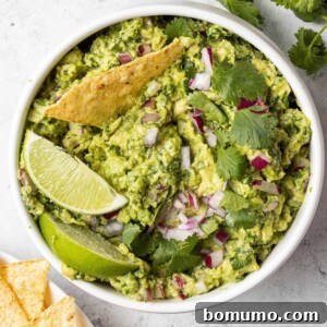 Round white bowl filled with guacamole, one chip dipped in, garnished with lime.