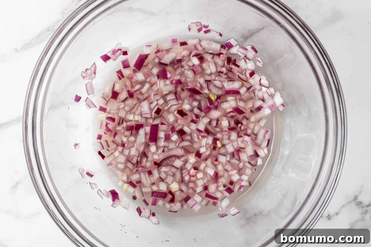 Crafting Perfect Guacamole 5 Finely diced red onions soaking in lime juice within a clear glass bowl, preparing them for guacamole.