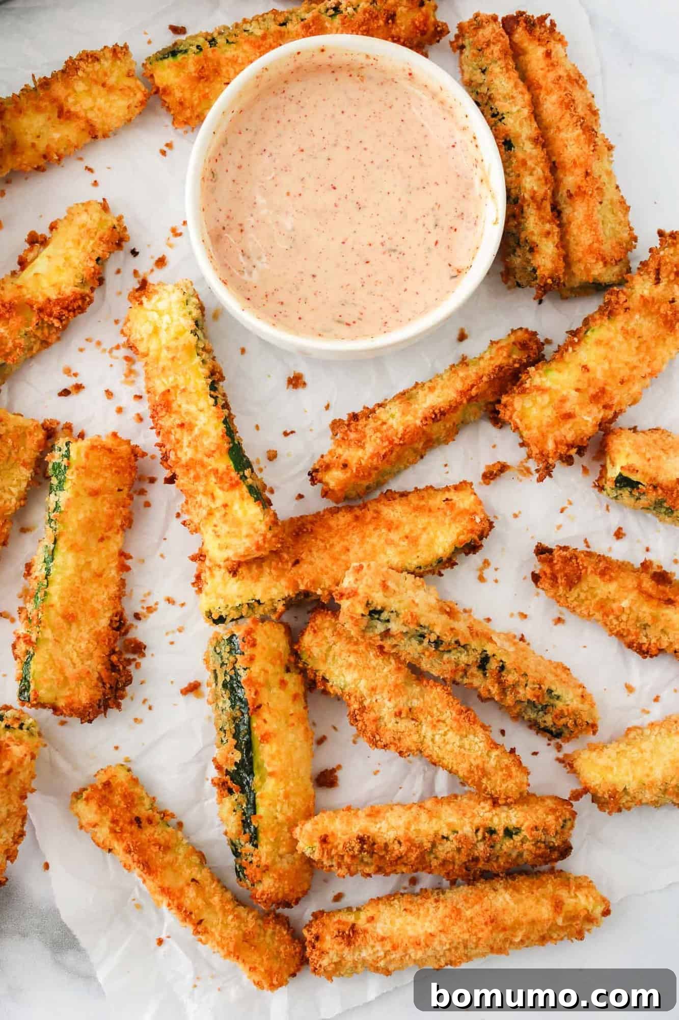 Crispy air fryer zucchini fries perfectly arranged on parchment paper, accompanied by a light red chili ranch dipping sauce.