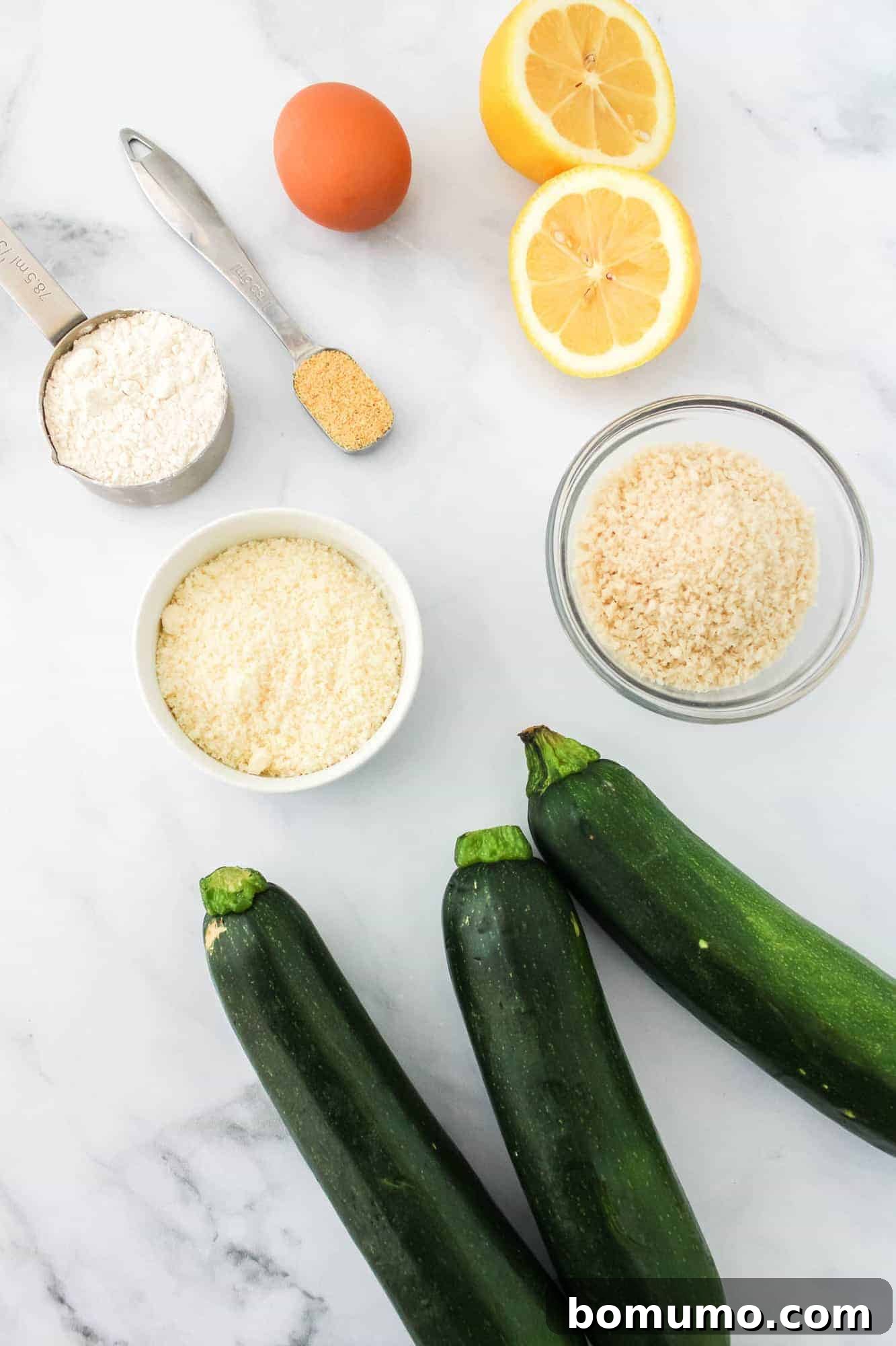 An overhead shot showcasing fresh zucchini, individual breading ingredients including flour, eggs, and panko mixture, alongside a halved lemon for zest.