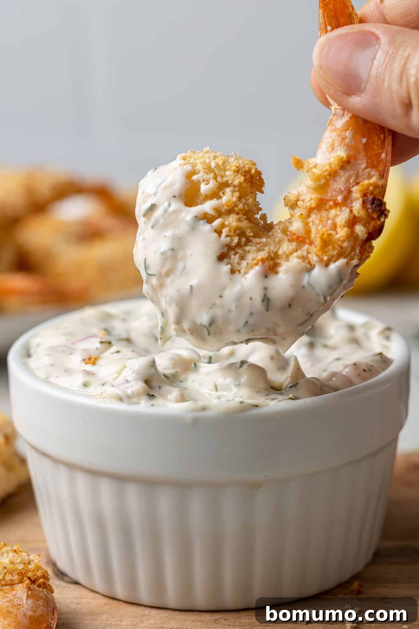 A hand holding a breaded shrimp, dipping it into a bowl of homemade tartar sauce, highlighting its creamy texture.