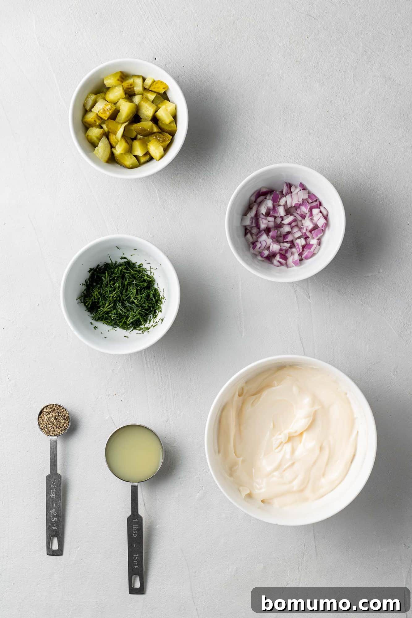 Overhead view of all individual ingredients needed for homemade tartar sauce, neatly arranged in small white bowls and with measuring spoons.