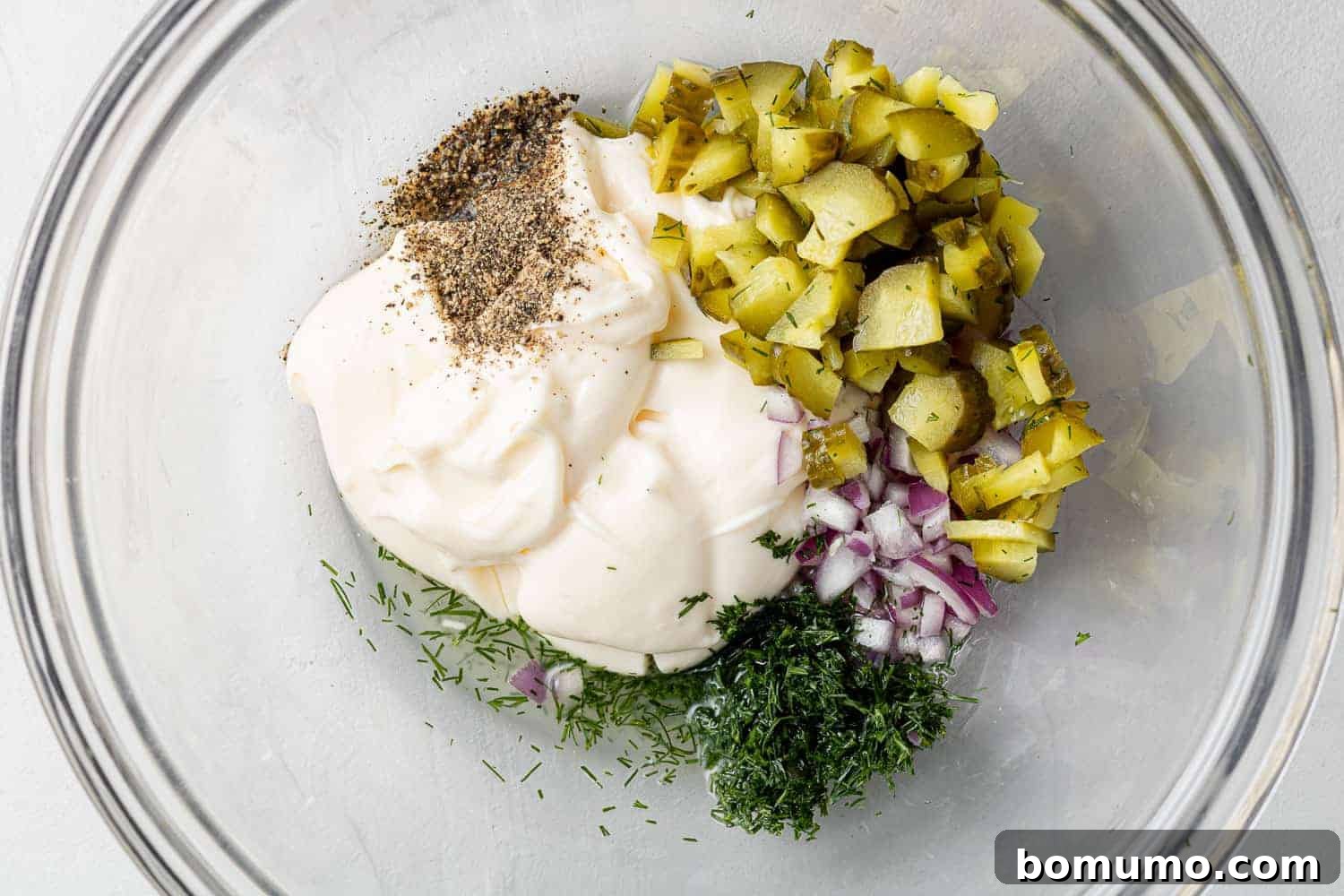 Ingredients for tartar sauce, including mayonnaise, chopped pickles, dill, onions, and pepper, unmixed in a bowl.