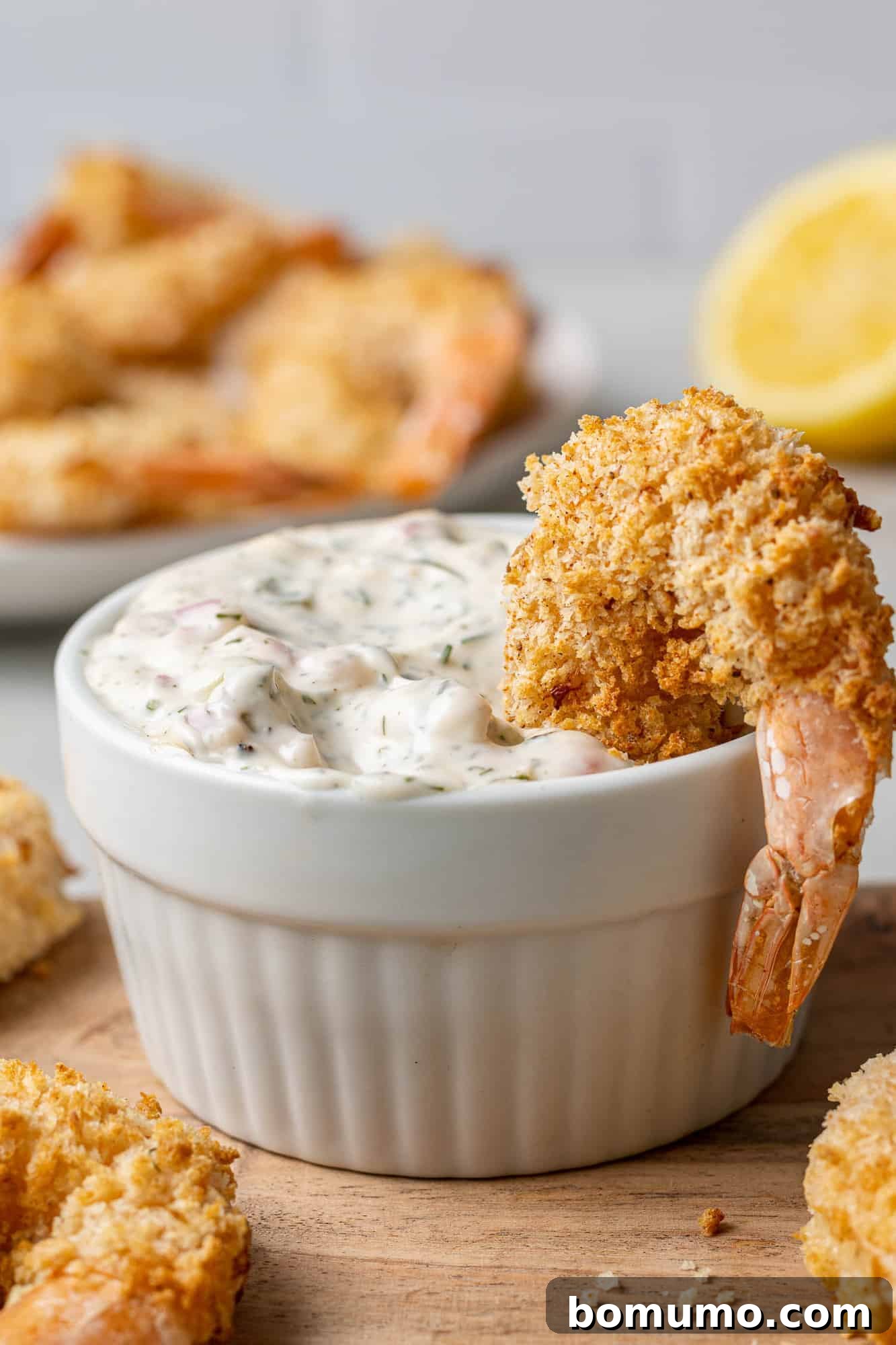 Crispy breaded shrimp positioned over the edge of a bowl of creamy homemade tartar sauce, ready for dipping.