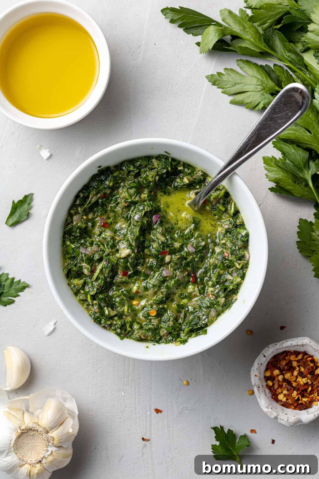 A top-down view of a white bowl filled with vibrant green chimichurri sauce, surrounded by its fresh ingredients like parsley, garlic, and red pepper flakes.