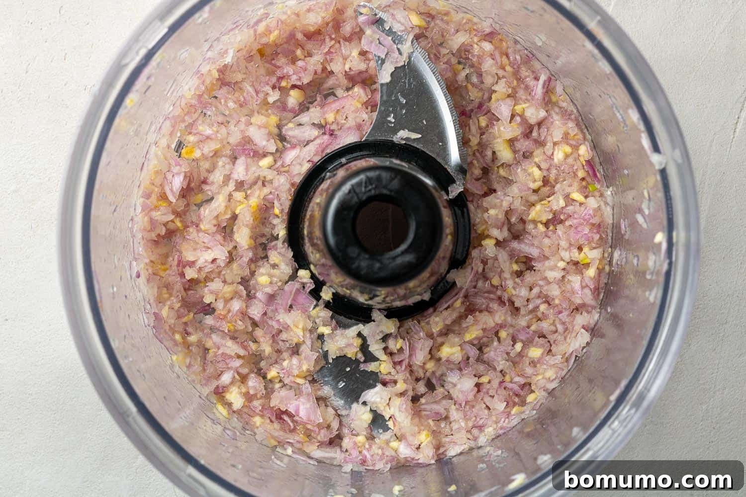Close-up overhead view of finely chopped shallots and garlic inside a food processor bowl, ready for further blending.