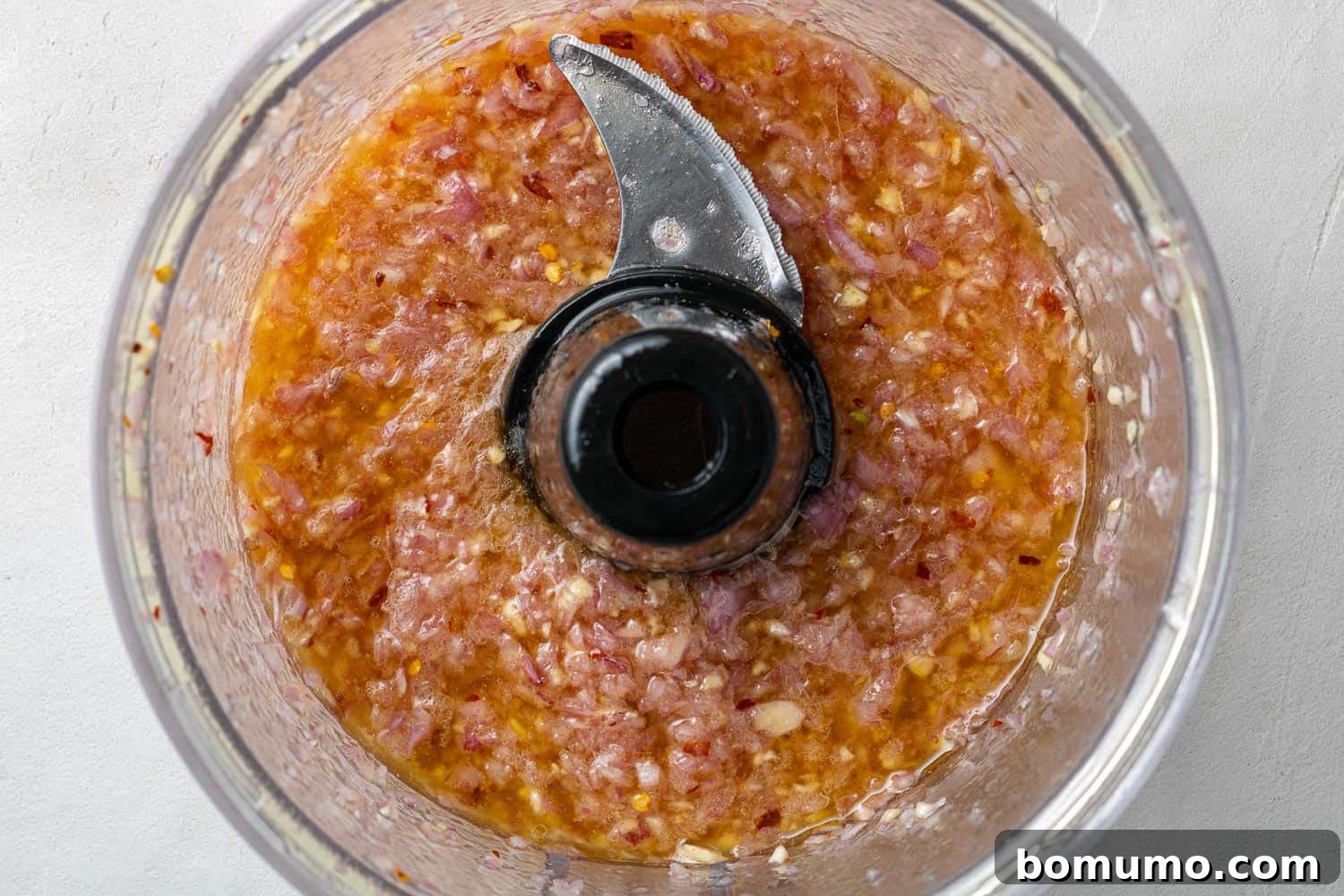 Overhead shot inside a food processor bowl, showing olive oil, red wine vinegar, and spices mixed with finely diced shallot and garlic.