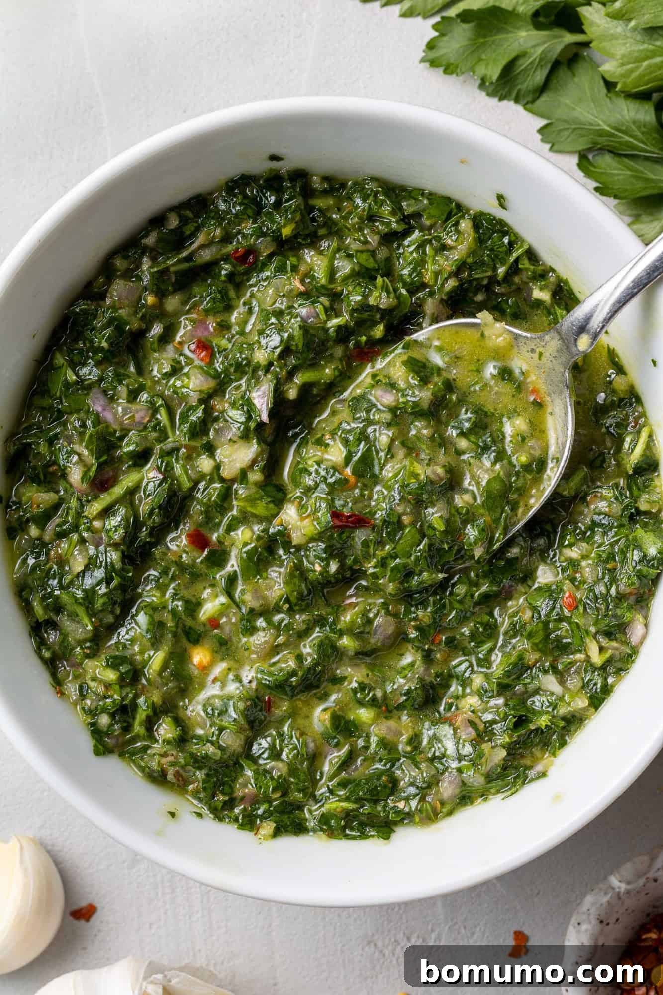 A close-up view of vibrant green chimichurri sauce in a white ceramic bowl, with a silver spoon scooping up a portion, highlighting its fresh texture.