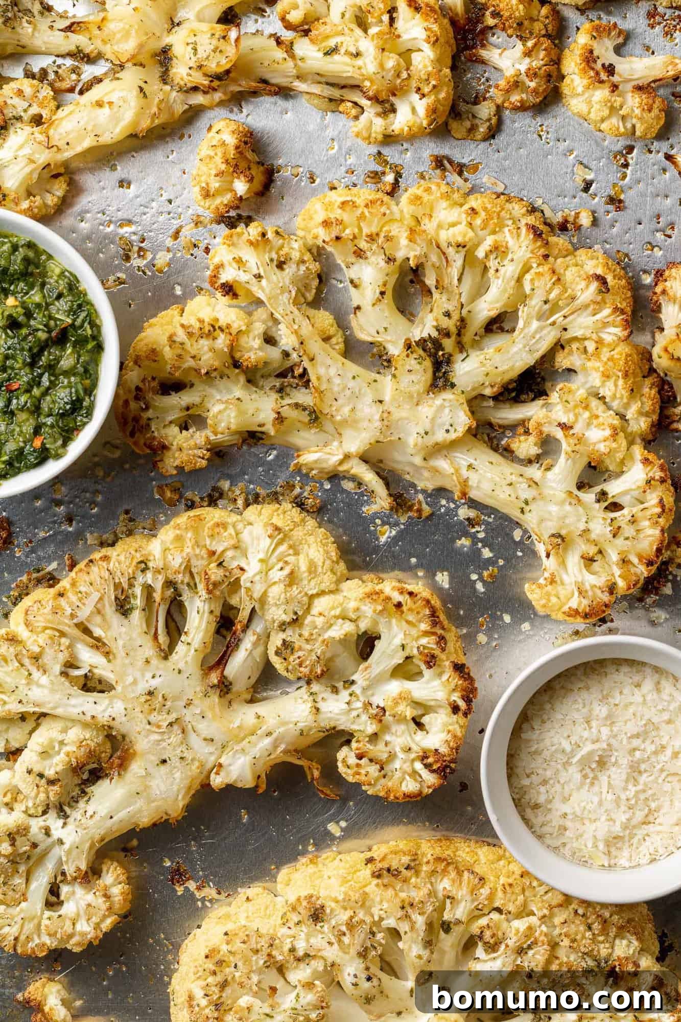 Beautifully roasted cauliflower steaks on a sheet pan, garnished with freshly grated parmesan and a vibrant dollop of green chimichurri sauce.
