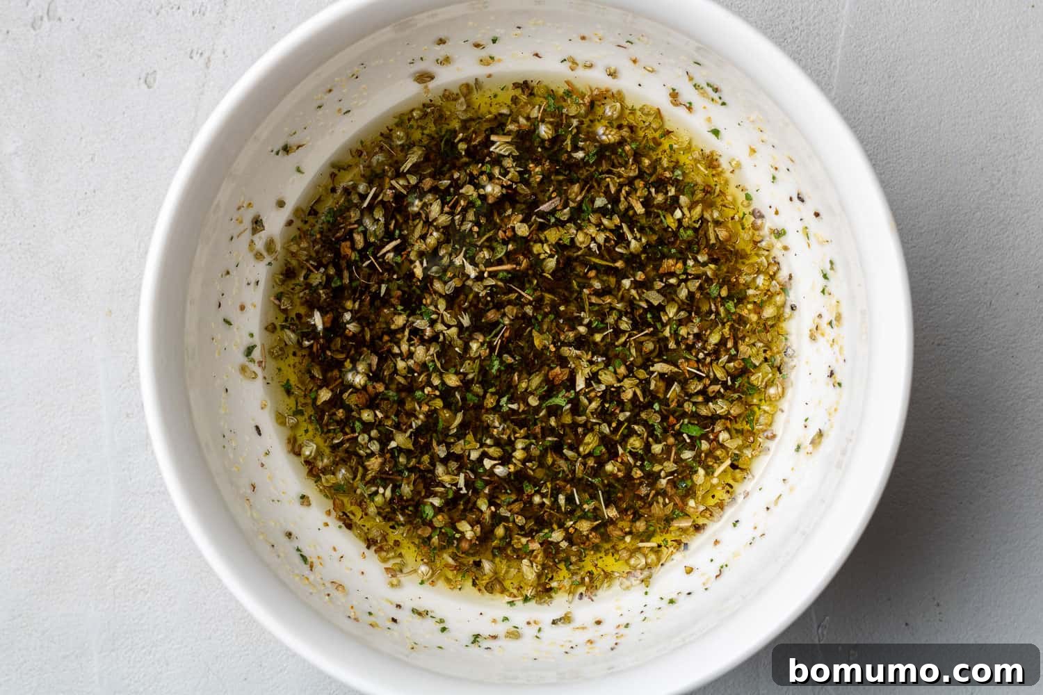 A small white bowl containing a mixture of olive oil and dried herbs and spices, ready for basting.
