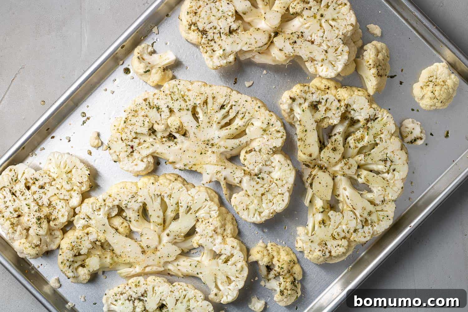Seasoned, yet uncooked, slices of cauliflower arranged on a sheet pan, waiting to be roasted.