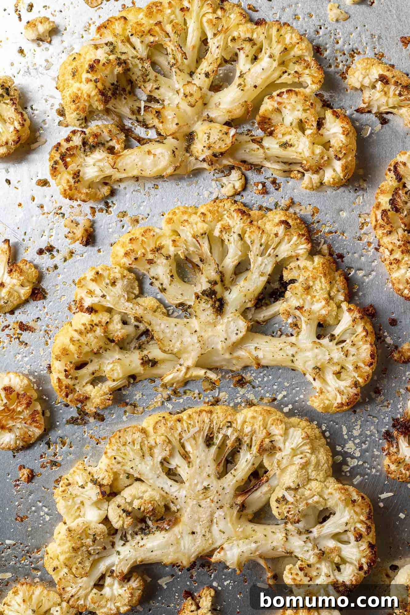 An overhead shot of three large, golden-brown cauliflower steaks arranged neatly on a silver sheet pan.