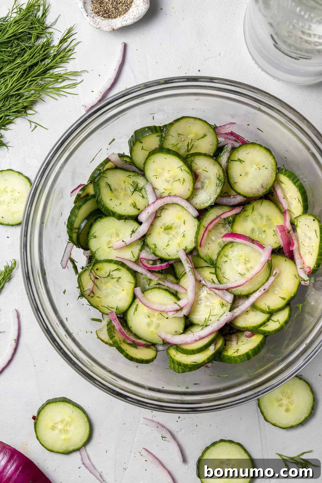 Overhead view of sliced cucumbers and onions in a clear glass bowl.