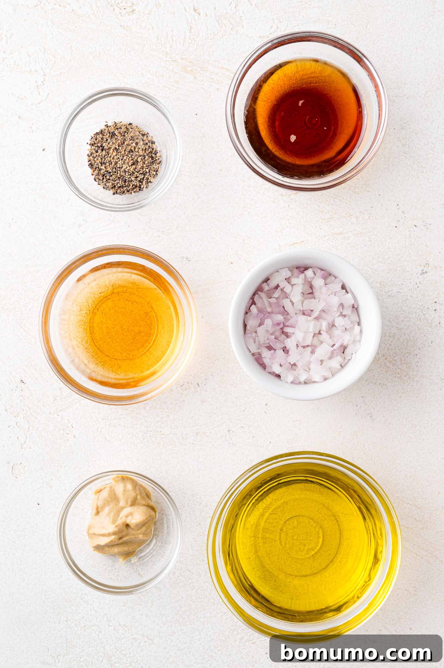 A selection of vinaigrette ingredients presented in small, elegant bowls on a light backdrop: ground black pepper, rich maple syrup, tangy apple cider vinegar, fragrant diced shallots, creamy Dijon mustard, and golden olive oil, all awaiting combination.