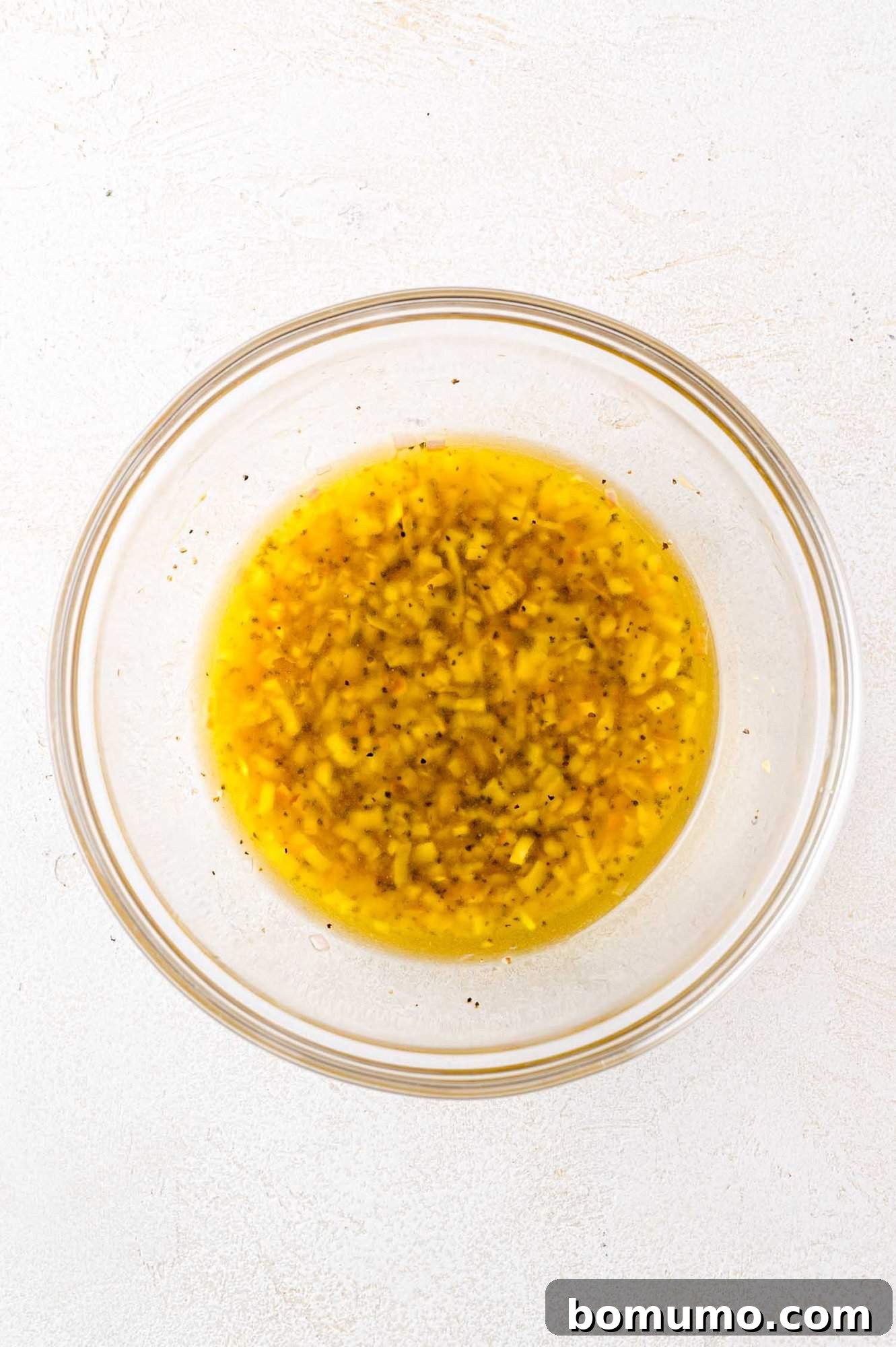 A clear glass bowl showcasing a freshly prepared, emulsified yellow vinaigrette, flecked with finely chopped herbs and spices, highlighting its vibrant texture and readiness to dress the salad.