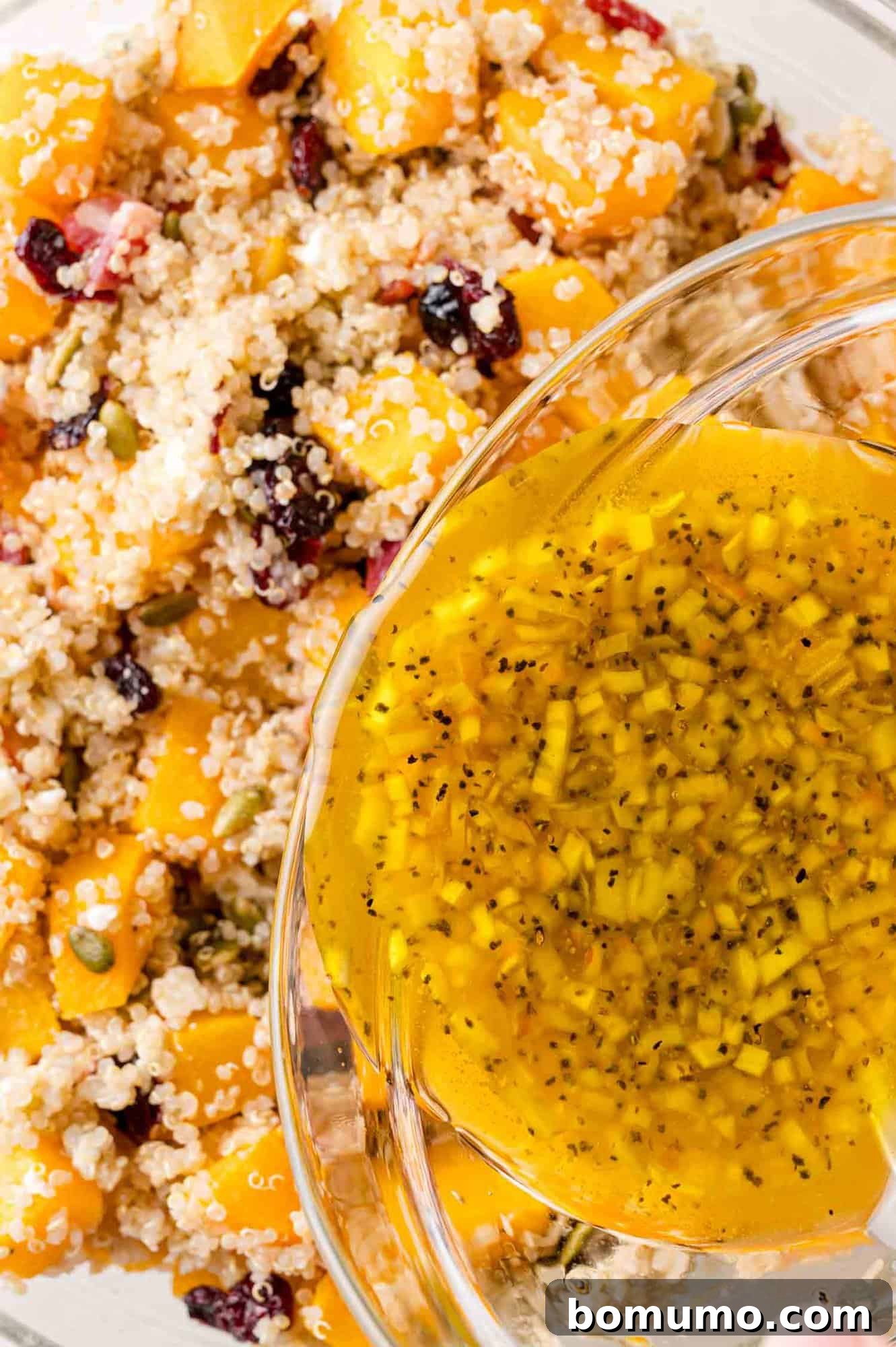 A close-up view of the vibrant quinoa salad, a harmonious mix of squash and cranberries, as a stream of the rich, golden dressing is poured over it, preparing it for a gentle toss.