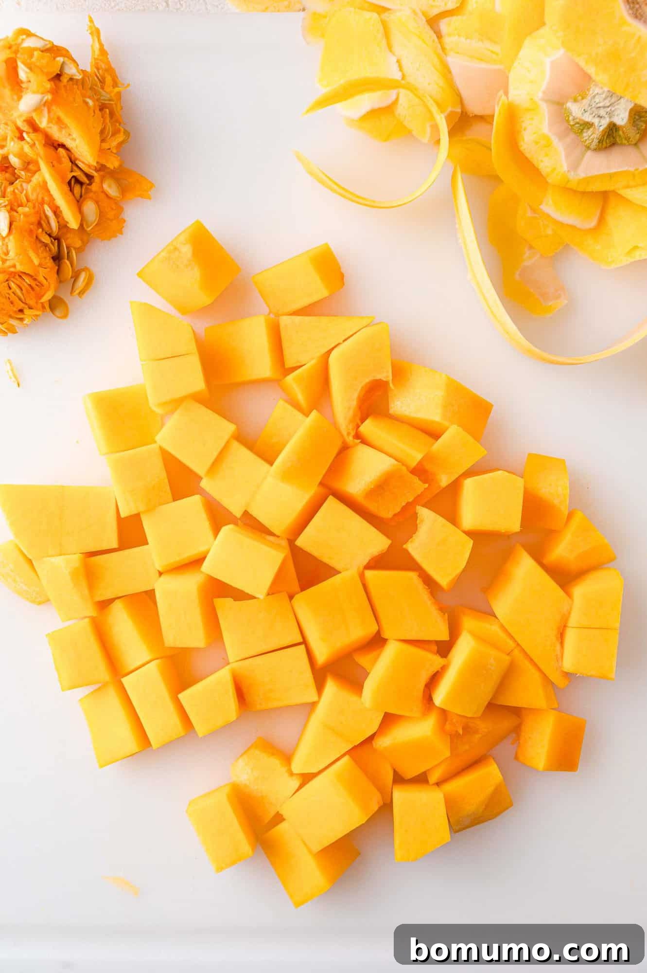 A white cutting board displaying neatly cubed butternut squash alongside its removed peelings and seeds, illustrating the preparation process for the salad.