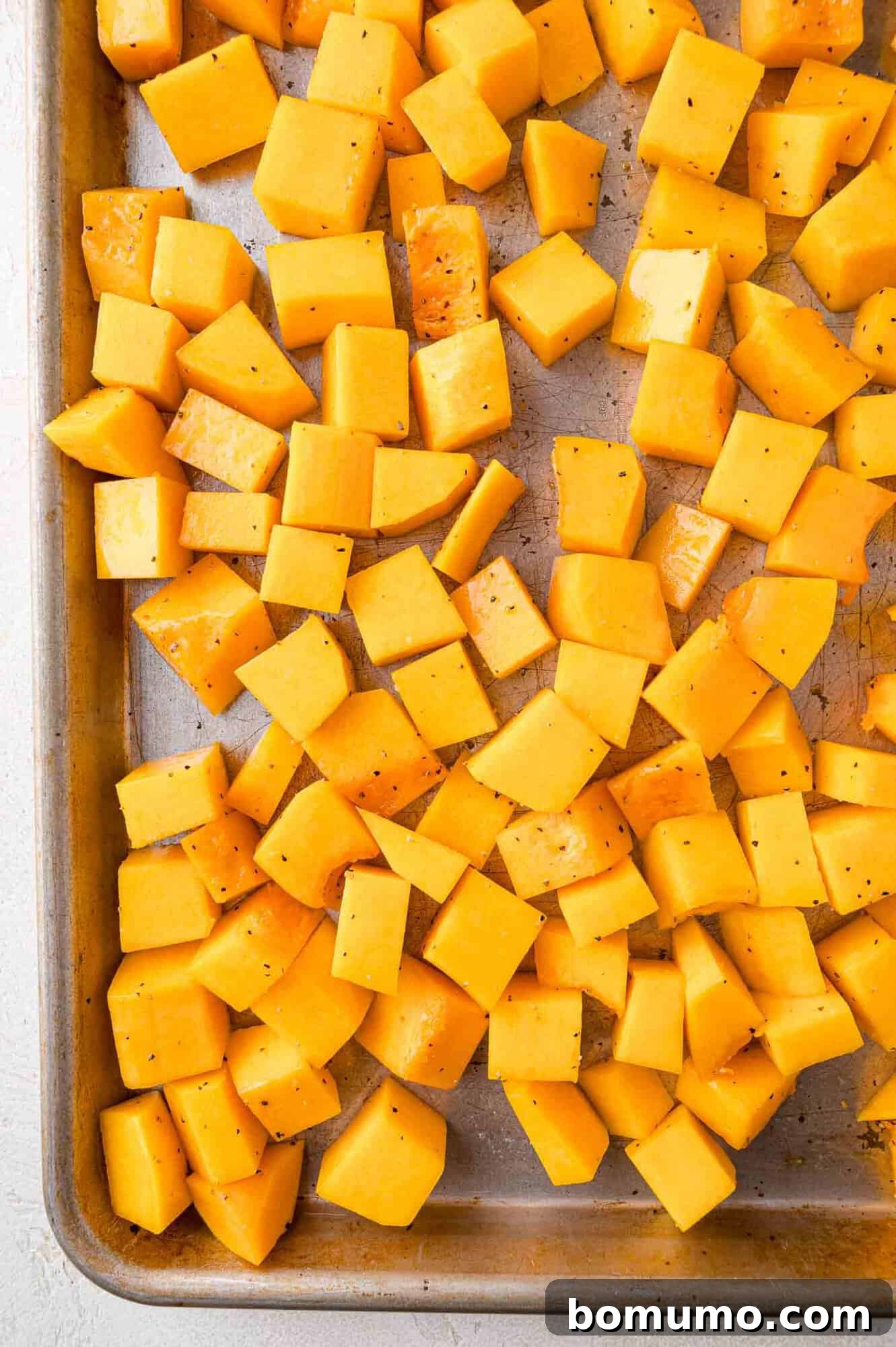 Cubed butternut squash, perfectly seasoned with freshly ground pepper, spread evenly on a baking sheet, poised for roasting to achieve a tender, caramelized finish.