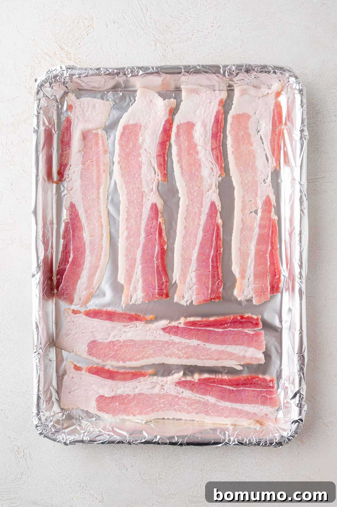 Raw bacon slices are neatly arranged in a single layer on a foil-lined baking sheet, prepared for effortless baking in the oven to achieve perfect crispness.
