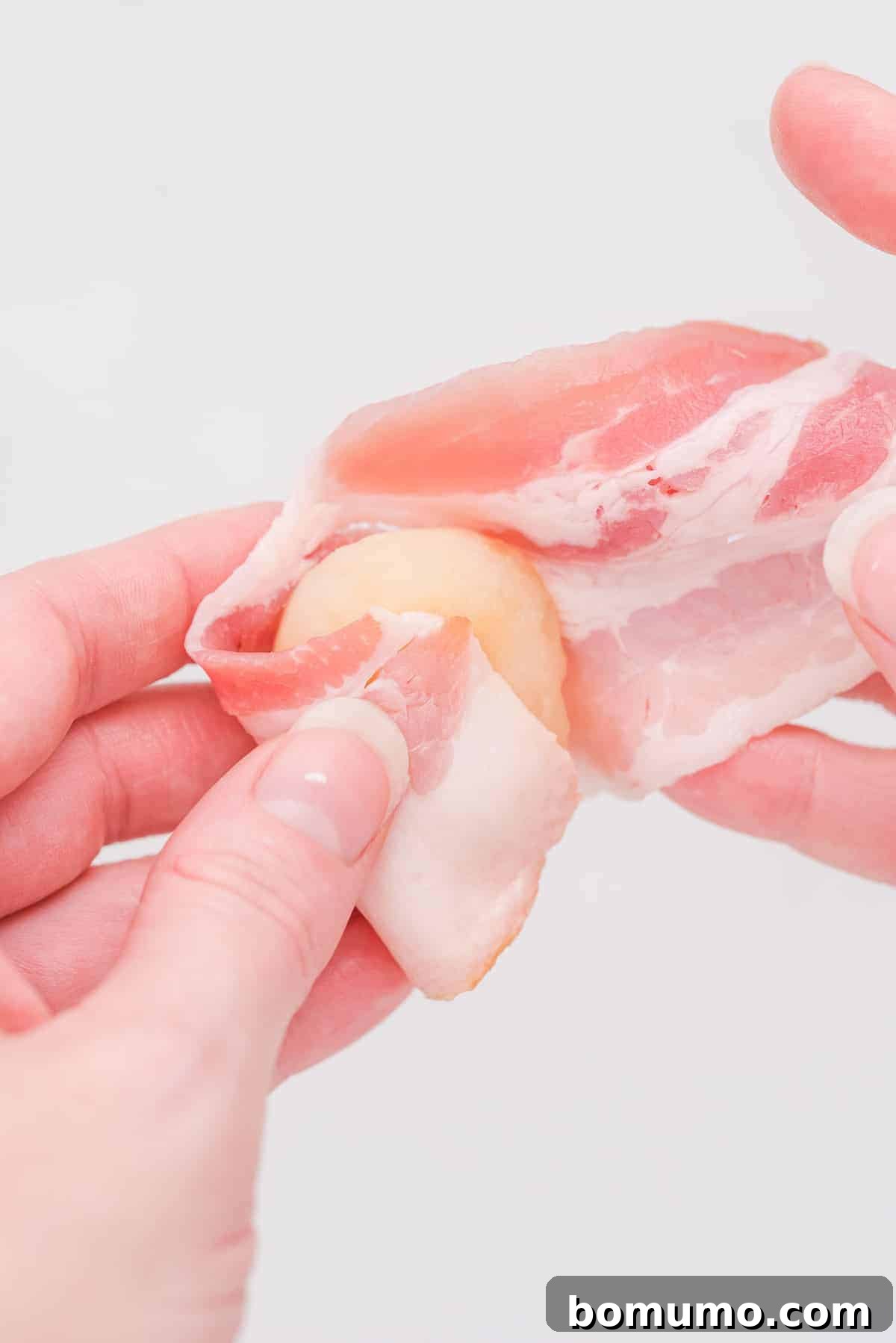 A visual guide showing how to expertly wrap a strip of bacon around a water chestnut, demonstrating the first step of preparation.