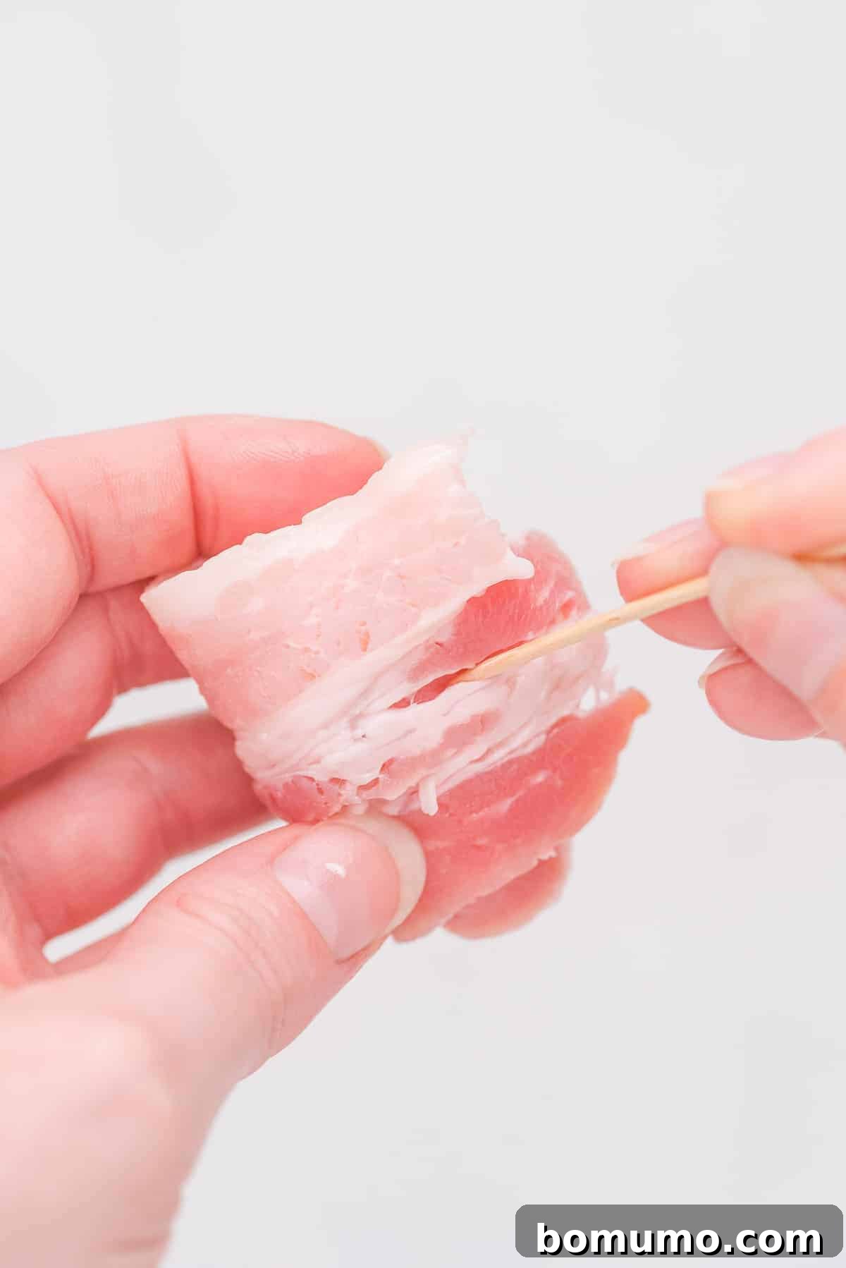 A wooden toothpick being inserted into an uncooked bacon-wrapped water chestnut, securing the bacon firmly in place before baking.