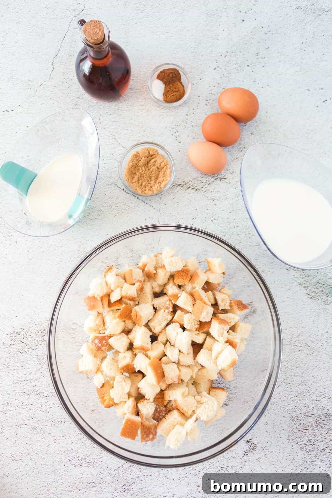 Overhead view of all fresh ingredients laid out for French toast muffins: bread, eggs, milk, cream, vanilla, and spices.