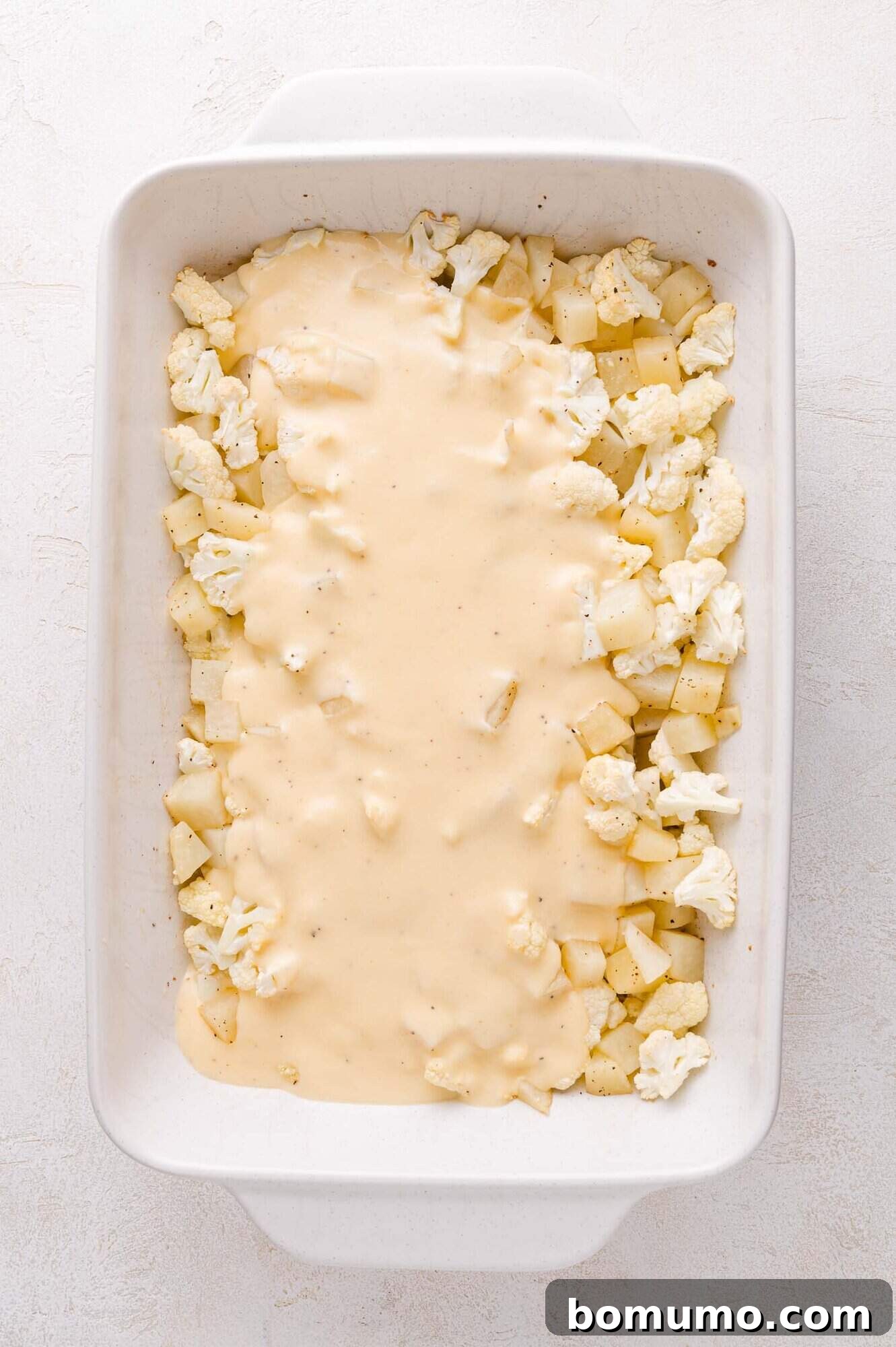 A white baking dish filled with roasted chopped cauliflower and potatoes, partially covered with a rich, creamy layer of cheese sauce, mid-assembly.