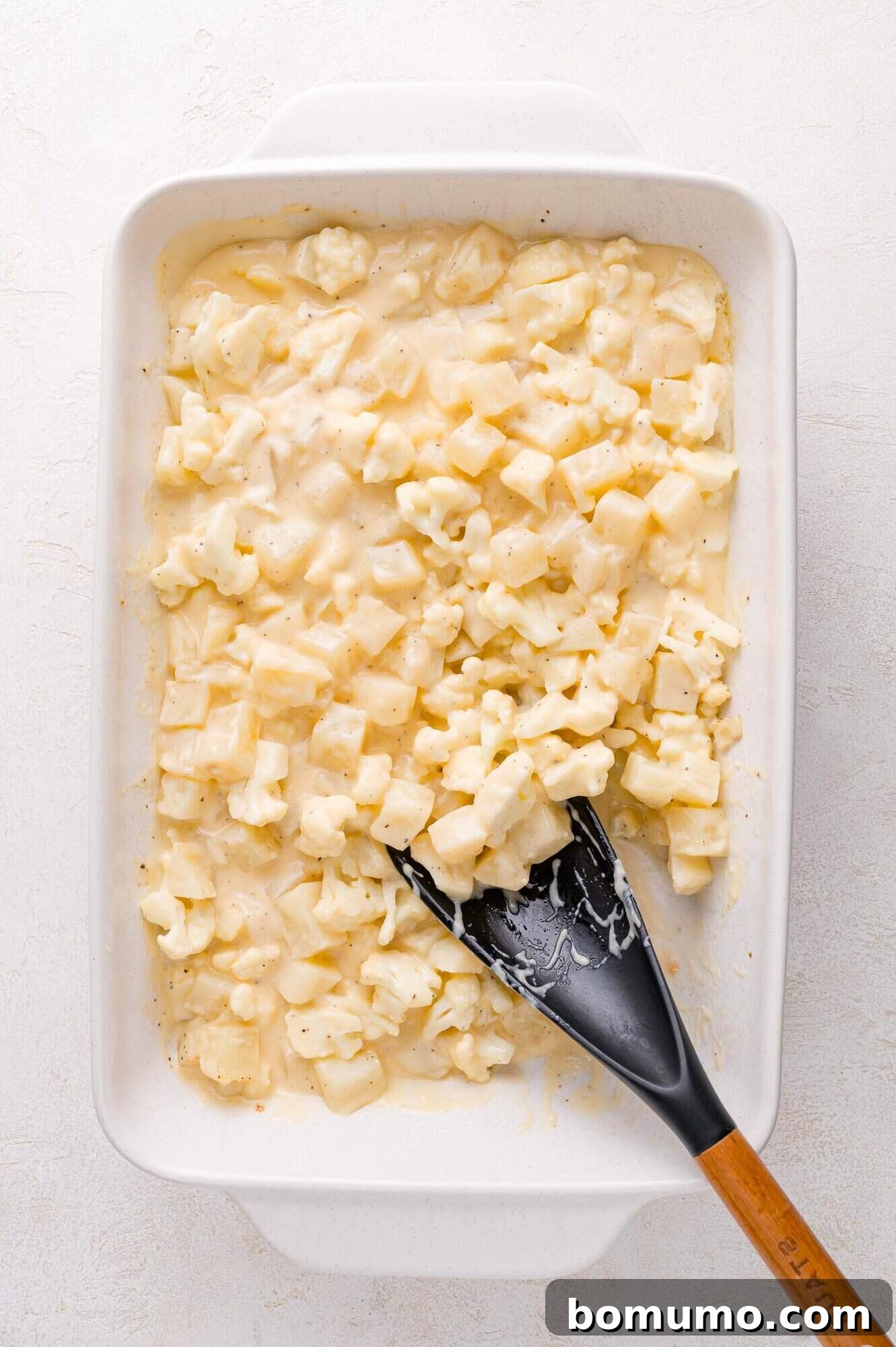 A baking dish filled with diced potatoes and cauliflower thoroughly mixed in a creamy cheese sauce, with a black spatula resting inside, ready for the topping.