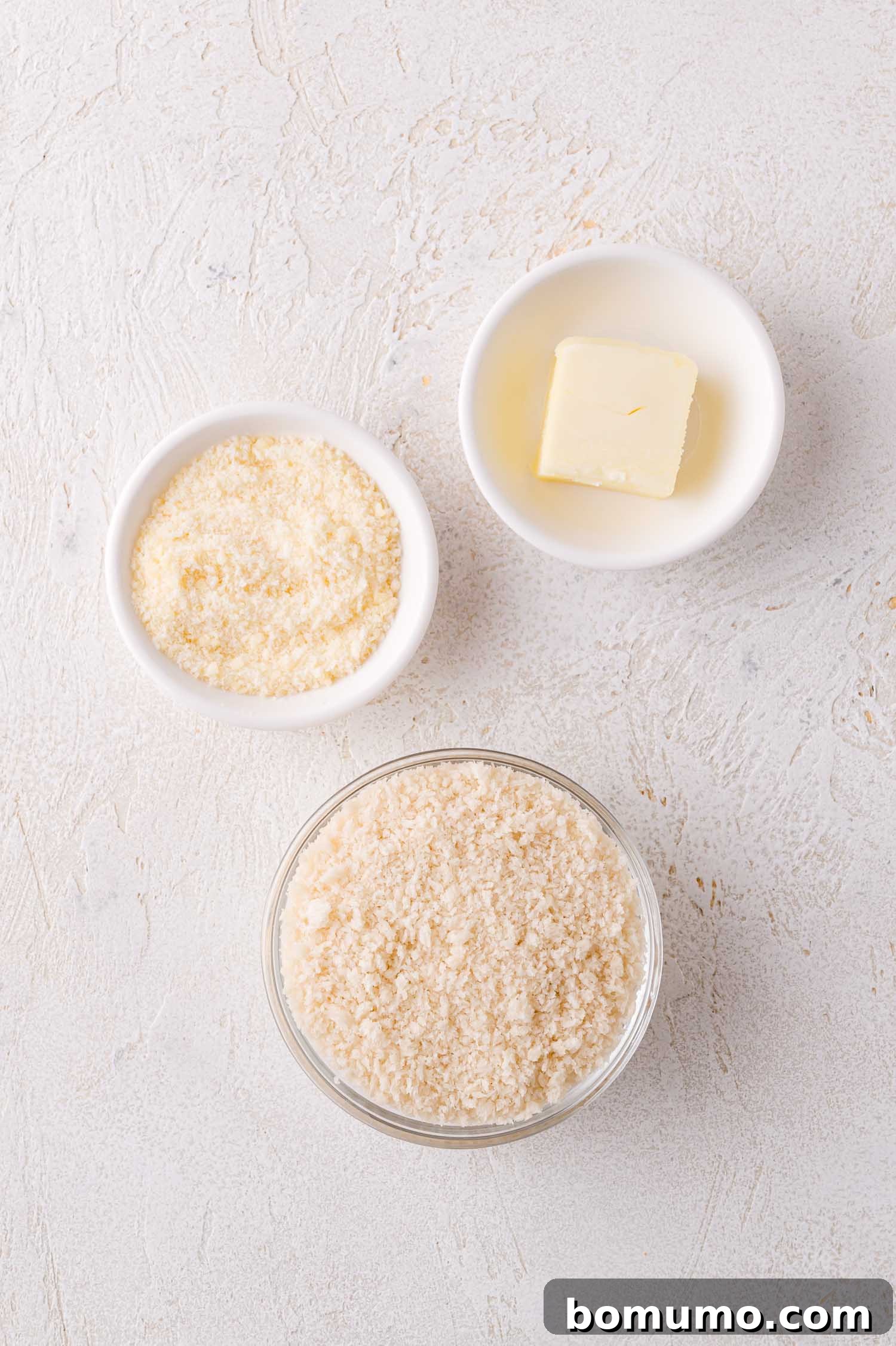 Three small bowls on a light surface: one with a pat of butter, one with grated Parmesan cheese, and one with panko breadcrumbs, ingredients for the topping.