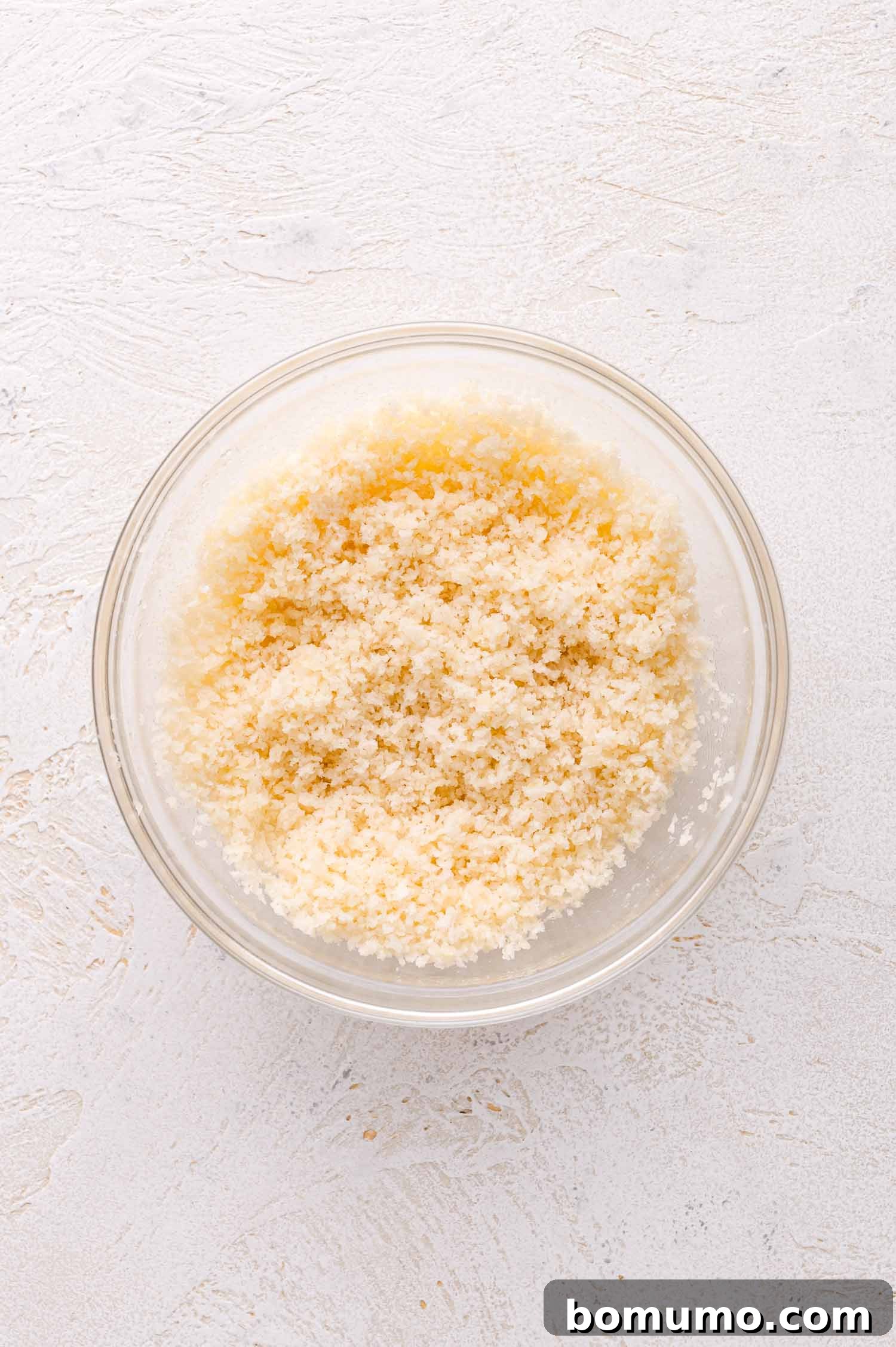 A clear glass bowl filled with light-colored panko breadcrumbs sits on a textured white surface, highlighting the coarse texture ideal for a crispy topping.