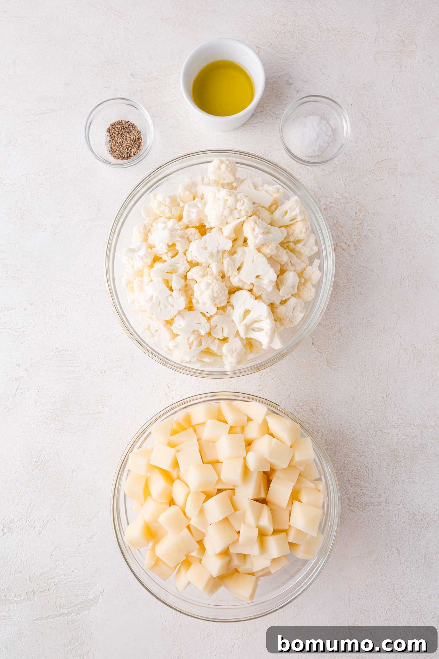 Two bowls containing cauliflower florets and diced potatoes, with small bowls of olive oil, salt, and pepper arranged above them on a light surface, ready for preparation.