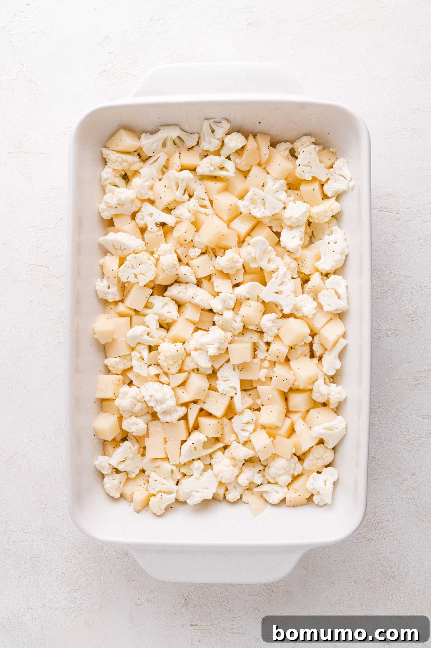 Chopped cauliflower and potatoes expertly arranged in a white rectangular baking dish on a light surface, seasoned and ready for roasting.