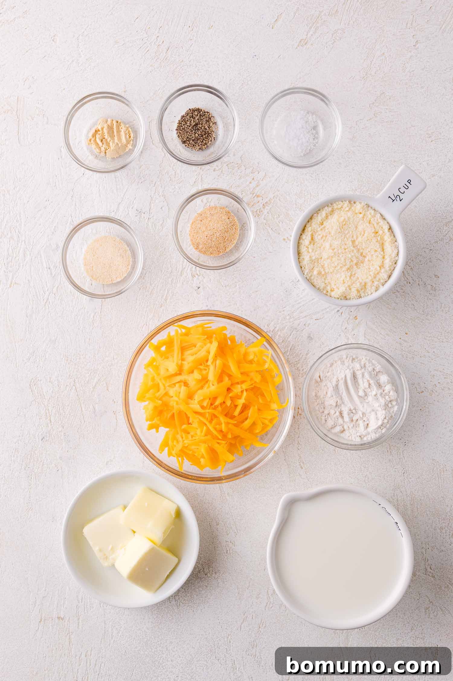 Assorted ingredients in small bowls, including shredded cheddar cheese, butter, milk, flour, bread crumbs, grated cheese, and various spices, arranged neatly on a white surface.