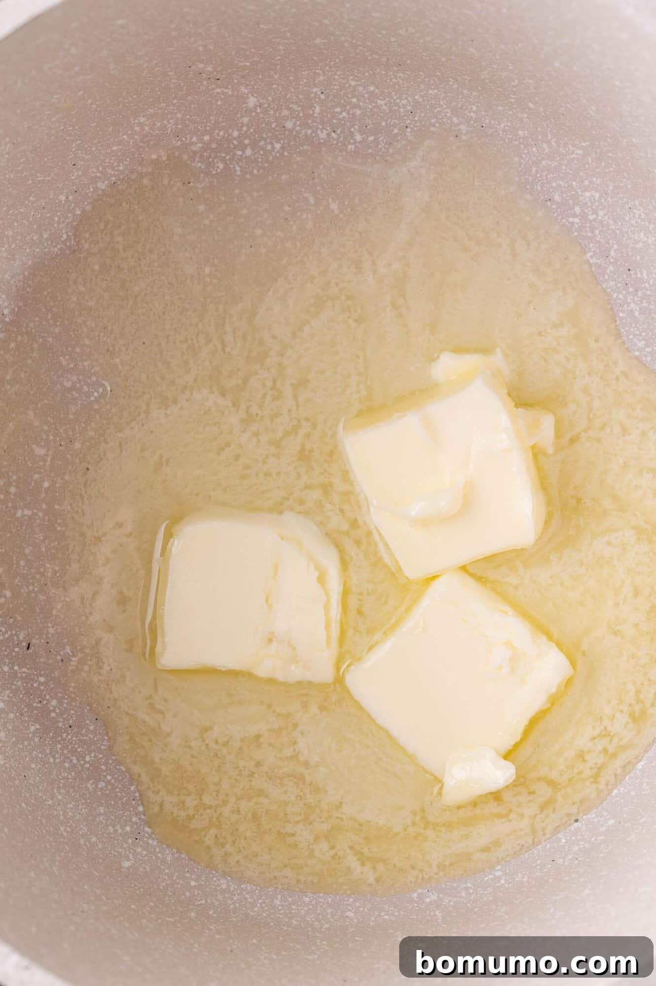 Three partially melted cubes of butter gently bubbling in a white saucepan on a stovetop, indicating the start of the cheese sauce.