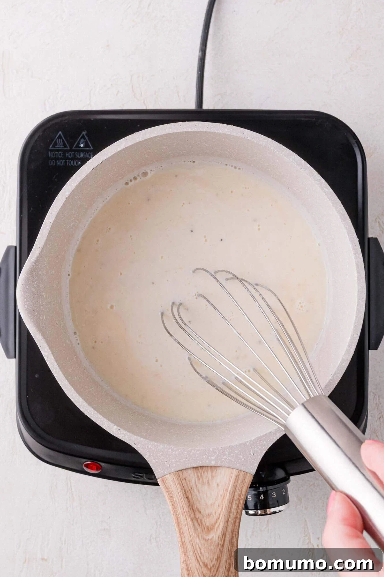 A hand expertly holds a whisk, stirring a creamy, bubbling sauce in a light-colored saucepan on a black electric hot plate, ensuring a smooth consistency.