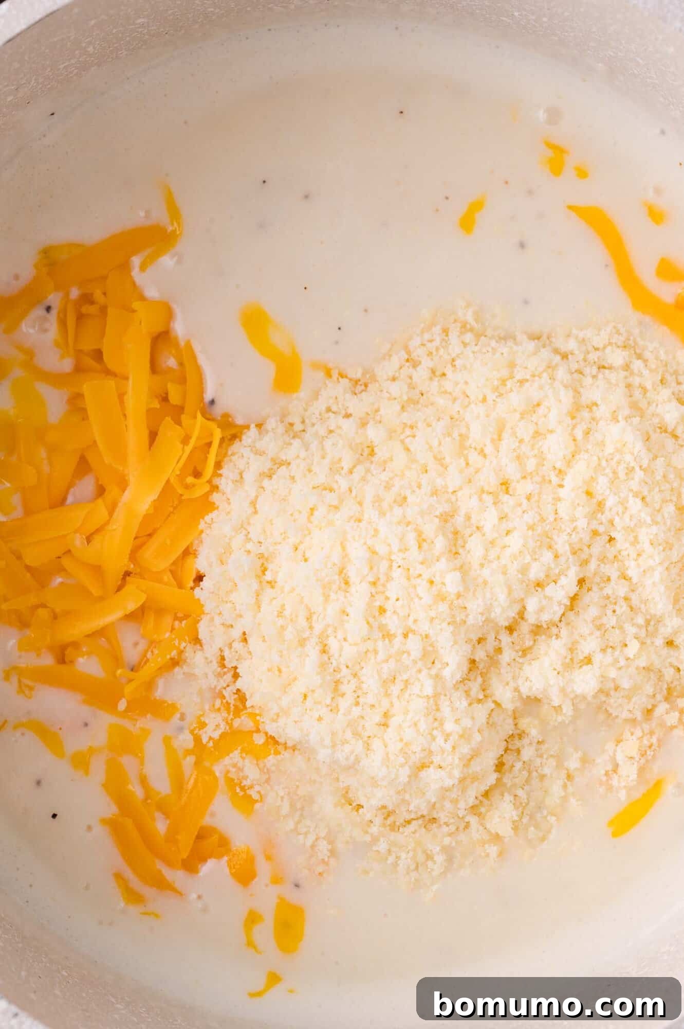 A generous amount of shredded cheddar cheese and grated Parmesan cheese being added to a creamy white sauce in a pot, ready to melt into a rich blend.