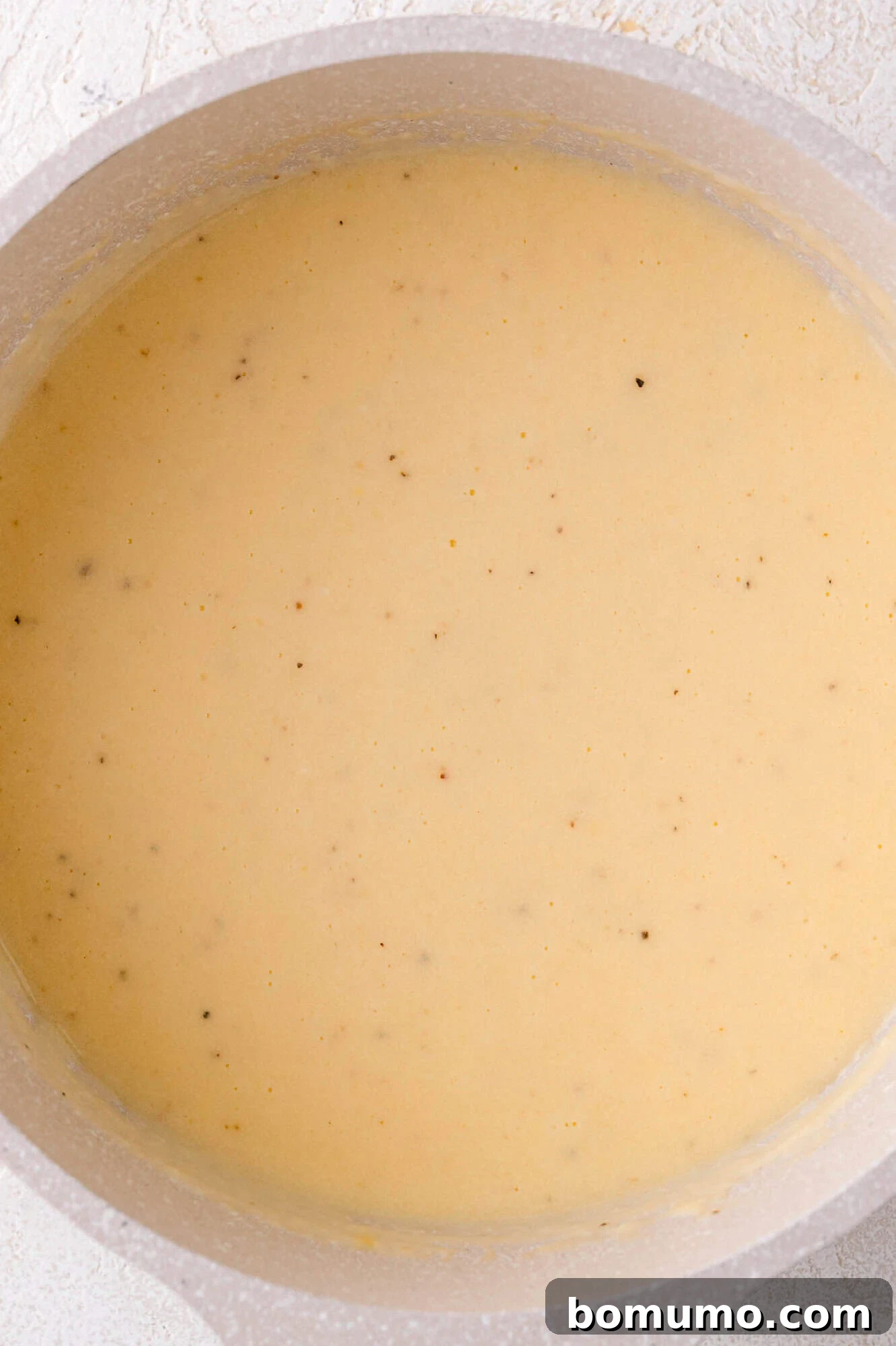 A pot filled with a perfectly smooth, creamy, light yellow cheese sauce, with subtle specks of seasoning visible, ready to be poured over vegetables.