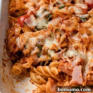 A delectable close-up of pizza pasta in a baking dish, brimming with pizza sauce, ham, cheese, tomatoes, and spinach, with a portion scooped out to highlight its rich layers.