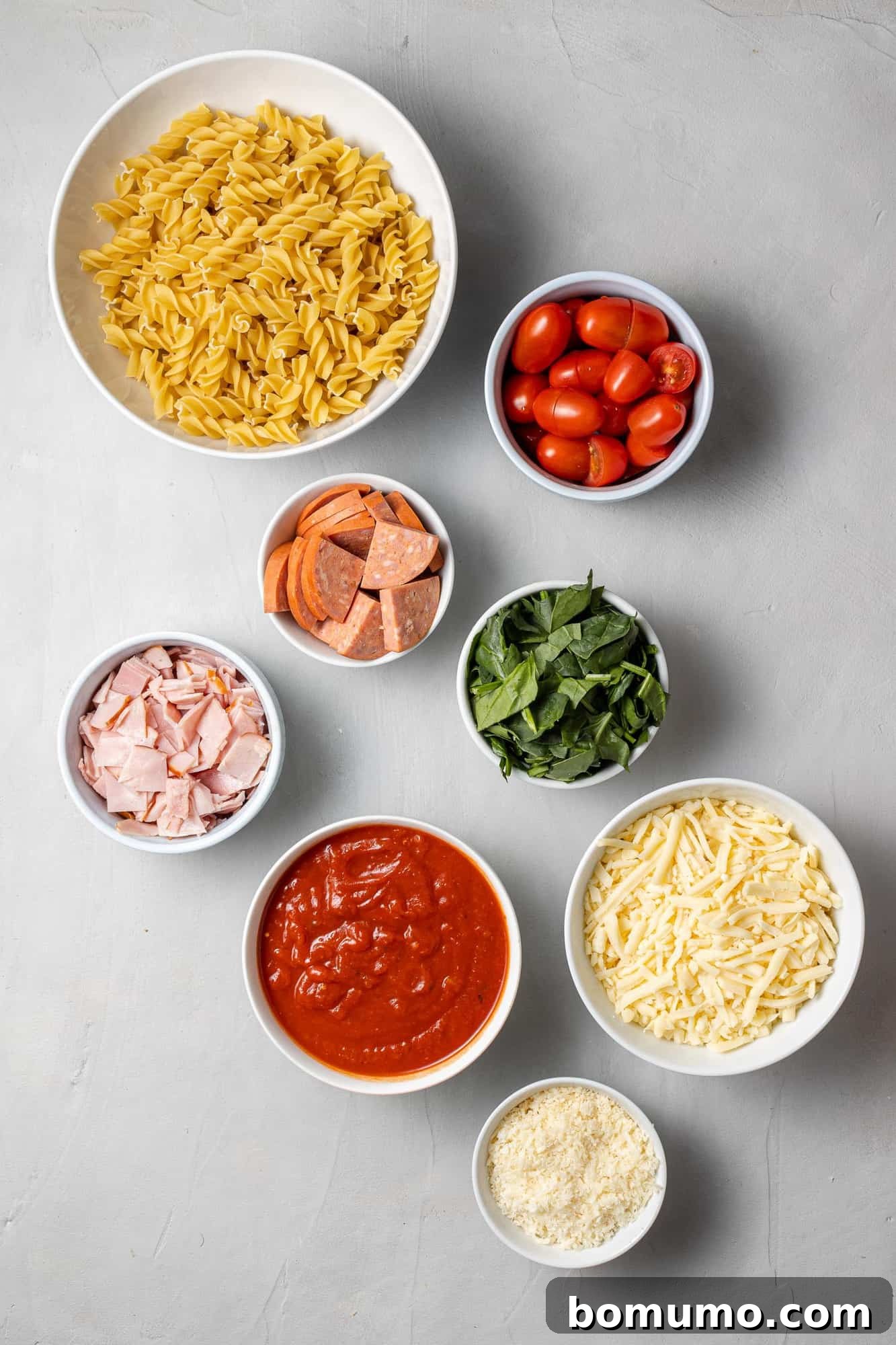 Overhead view of all the fresh and prepped ingredients laid out on a clean surface, including cooked pasta, tomatoes, ham, and pepperoni, ready for assembly.