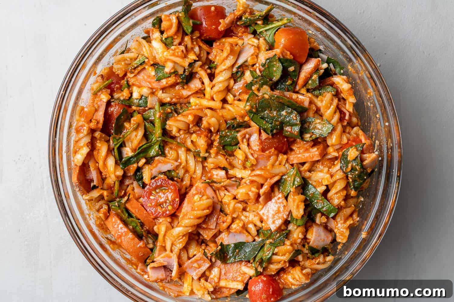 Close-up view of cooked pasta expertly mixed with vibrant red pizza sauce and a medley of savory toppings, all within a clear glass mixing bowl, awaiting its final bake.