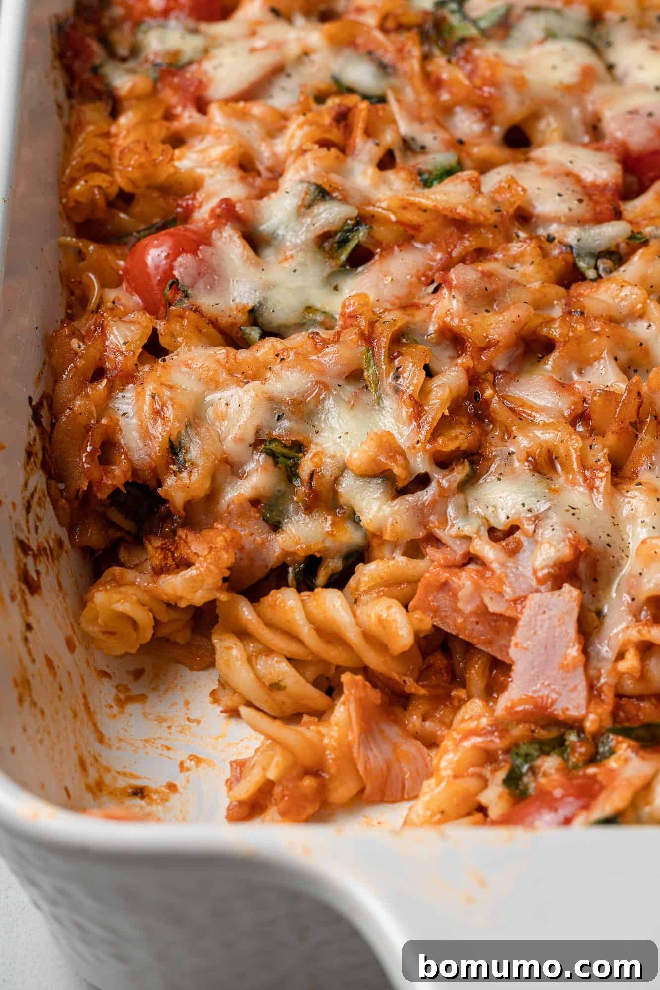 A close-up, overhead view of the perfectly baked pizza pasta casserole, with a generous scoop removed to reveal the layers of pasta, sauce, melted cheese, and colorful toppings, indicating it's ready to be devoured.