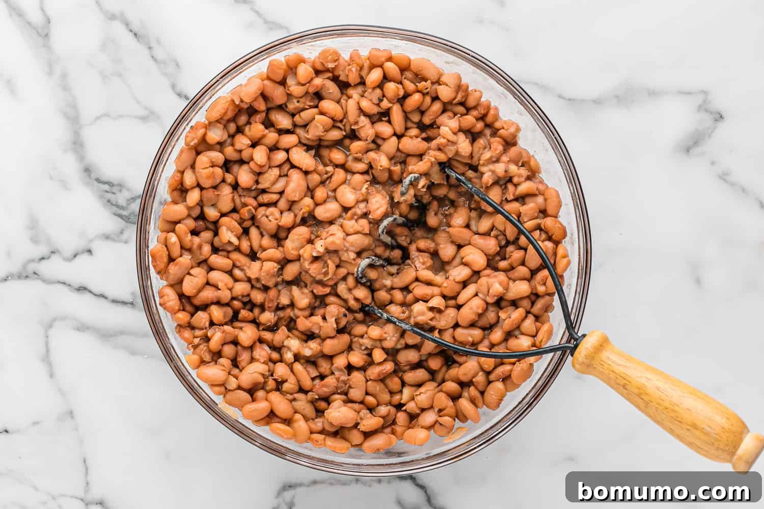 A close-up shot of tender pinto beans being mashed with a potato masher directly in the slow cooker, steam gently rising.