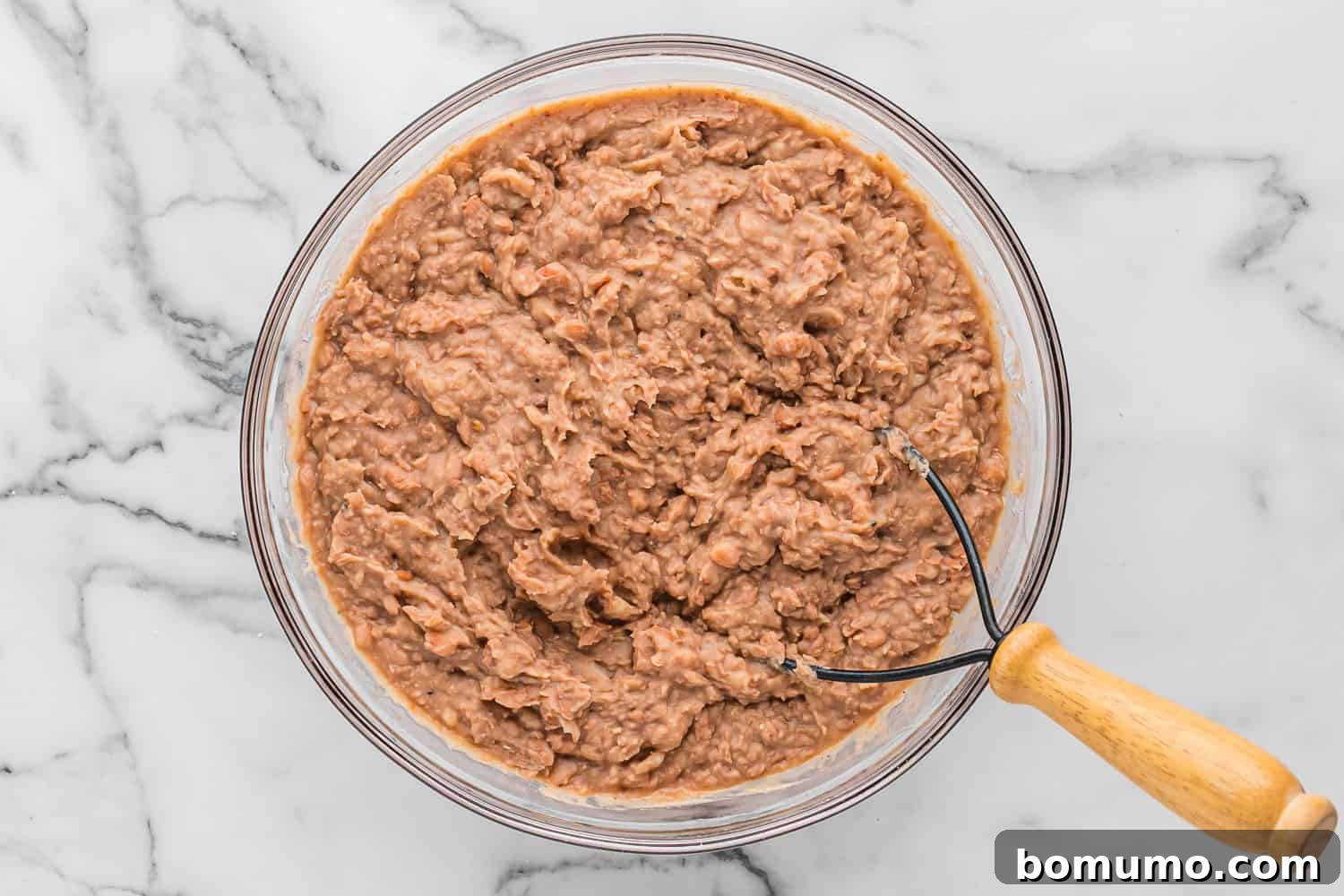 A close-up of beautifully mashed refried beans with a potato masher resting in the bowl, showcasing their smooth, creamy texture.