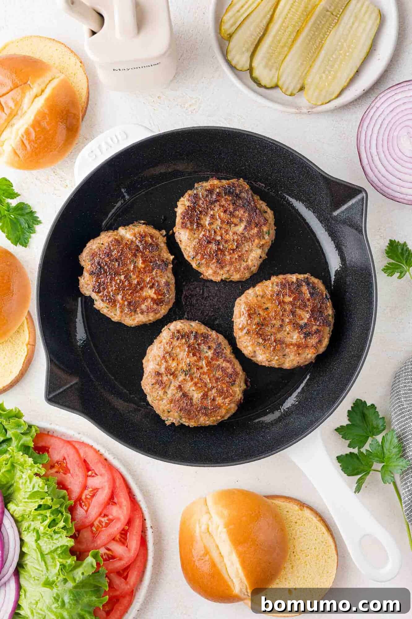 Four perfectly cooked turkey burger patties resting in a white-handled skillet, surrounded by an array of fresh burger accompaniments: soft burger buns, crisp lettuce, juicy tomato slices, tangy pickles, and thinly sliced red onion, all artfully arranged on a bright, inviting surface.
