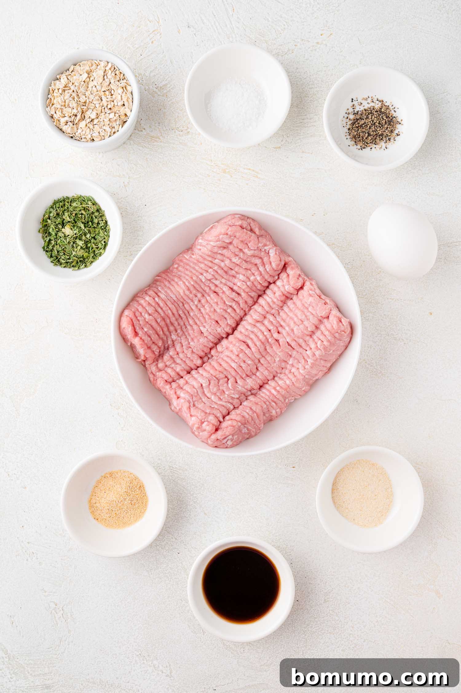 A large mixing bowl filled with raw ground meat, surrounded by smaller bowls containing various dry ingredients such as oats, salt, pepper, and other seasonings, all arranged on a light-colored surface, ready for blending.