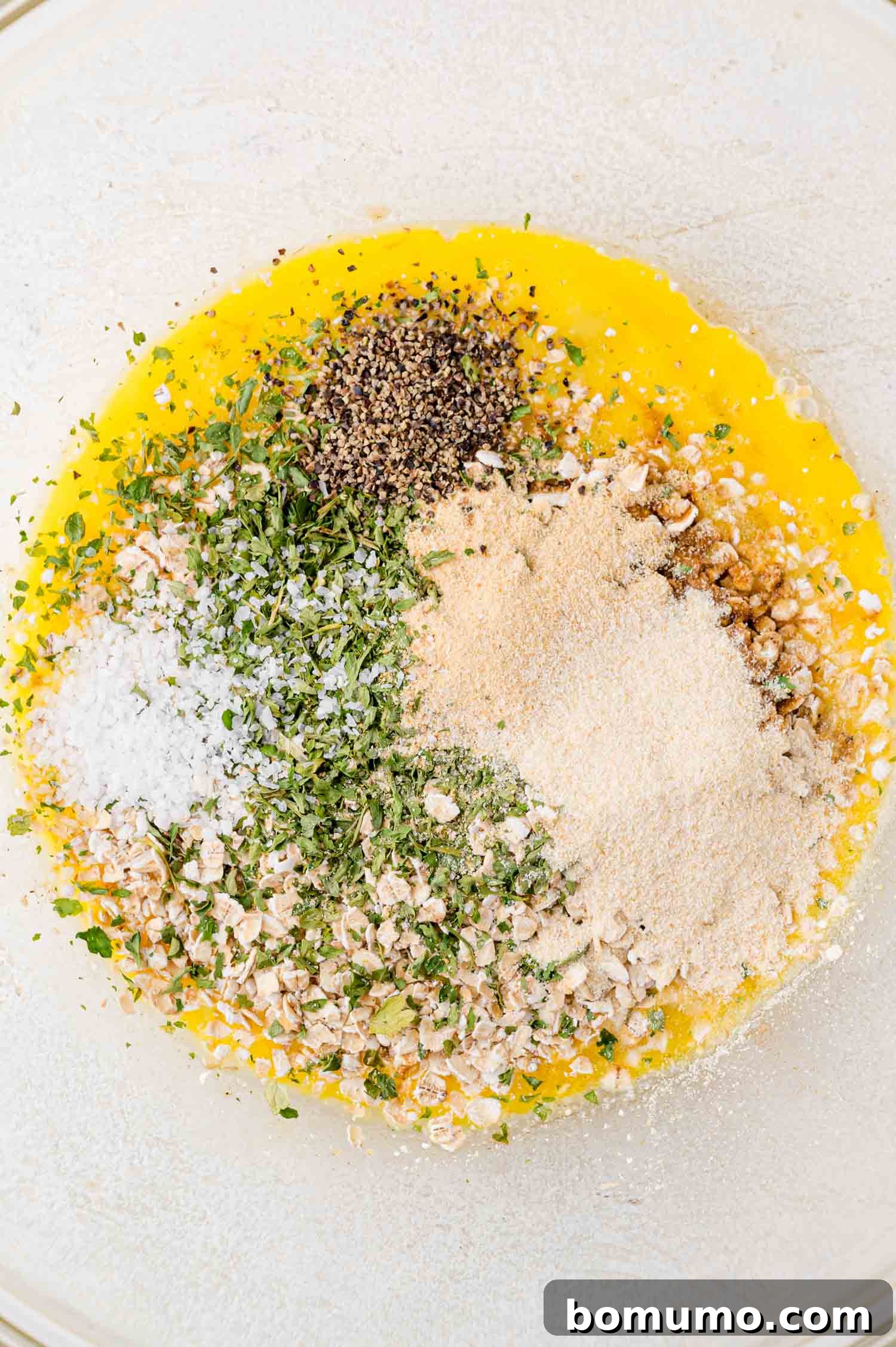 A glass bowl holds a vibrant mix of ingredients including beaten eggs, fresh herbs, and spices, surrounded by bowls of grated cheese and breadcrumbs, all set for a delicious recipe.