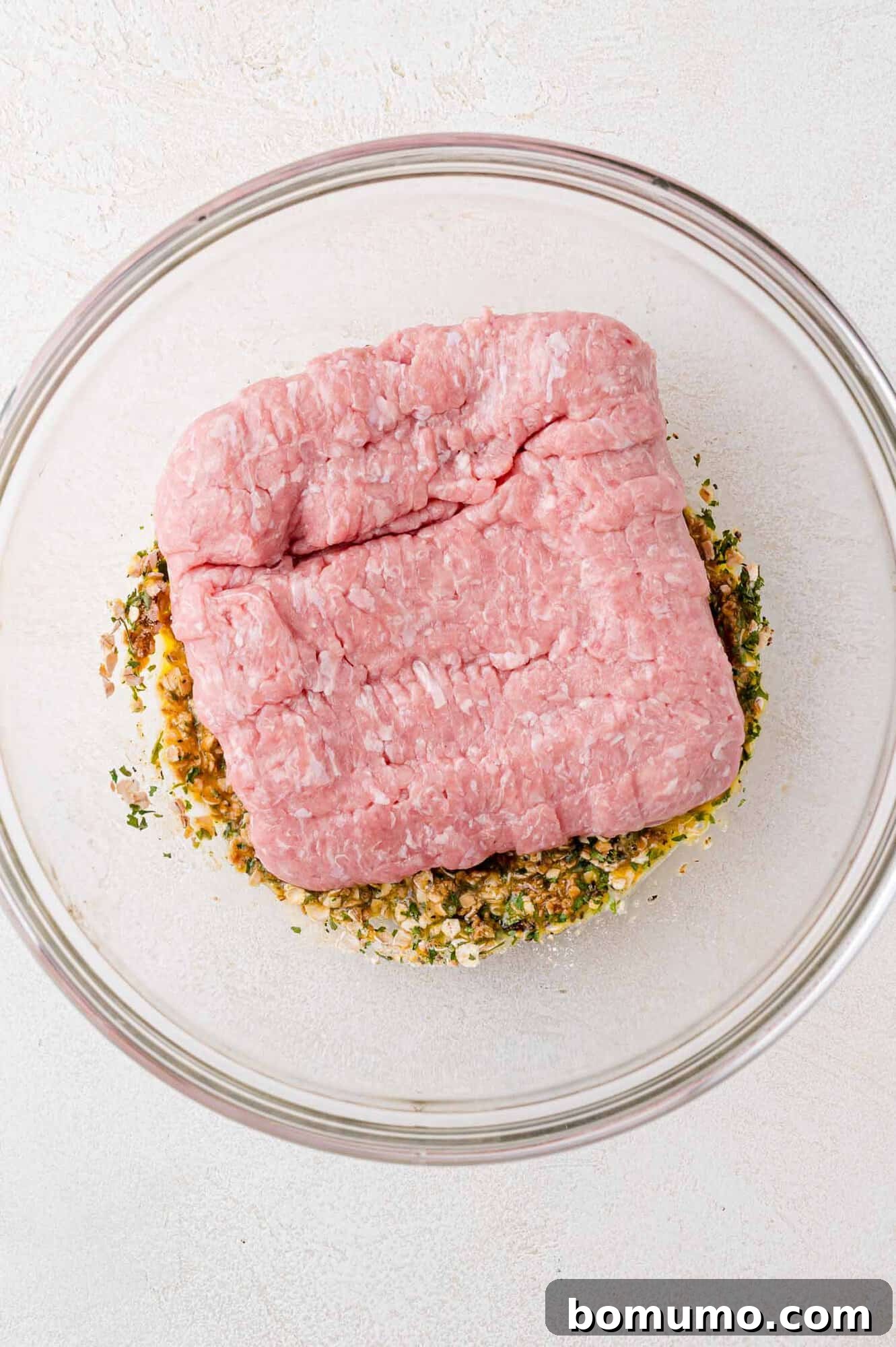 Raw ground turkey meticulously placed atop a savory blend of eggs and fresh herbs within a clear glass bowl, resting on a clean white surface.